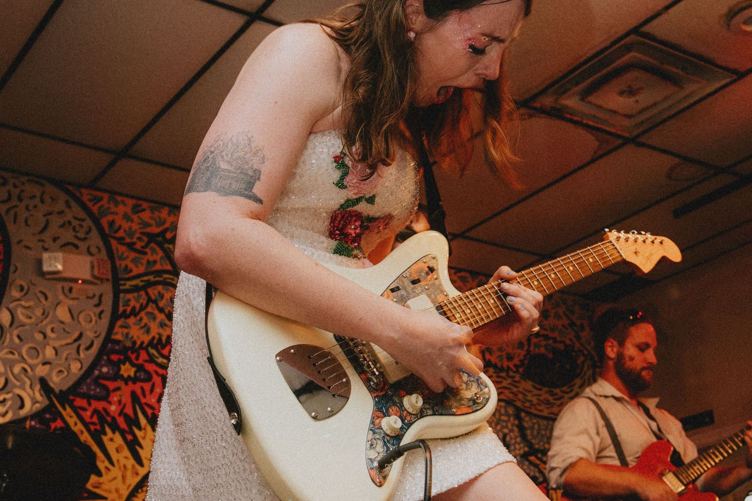 Woman in floral dress plays white Fender guitar on stage while man with beard plays guitar in background.