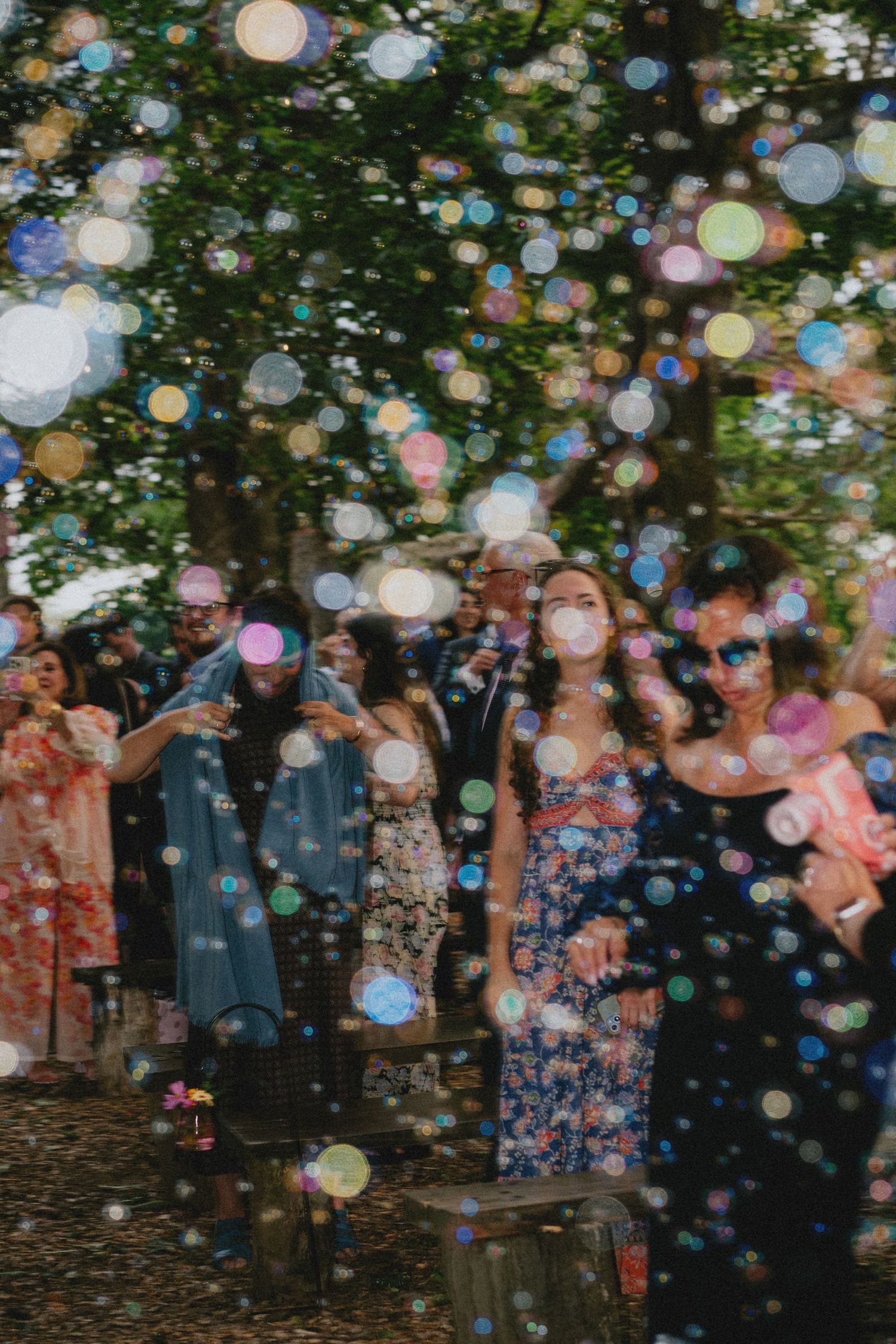 Guests celebrate outdoors surrounded by colorful floating bubbles at a wedding reception under lush green trees.