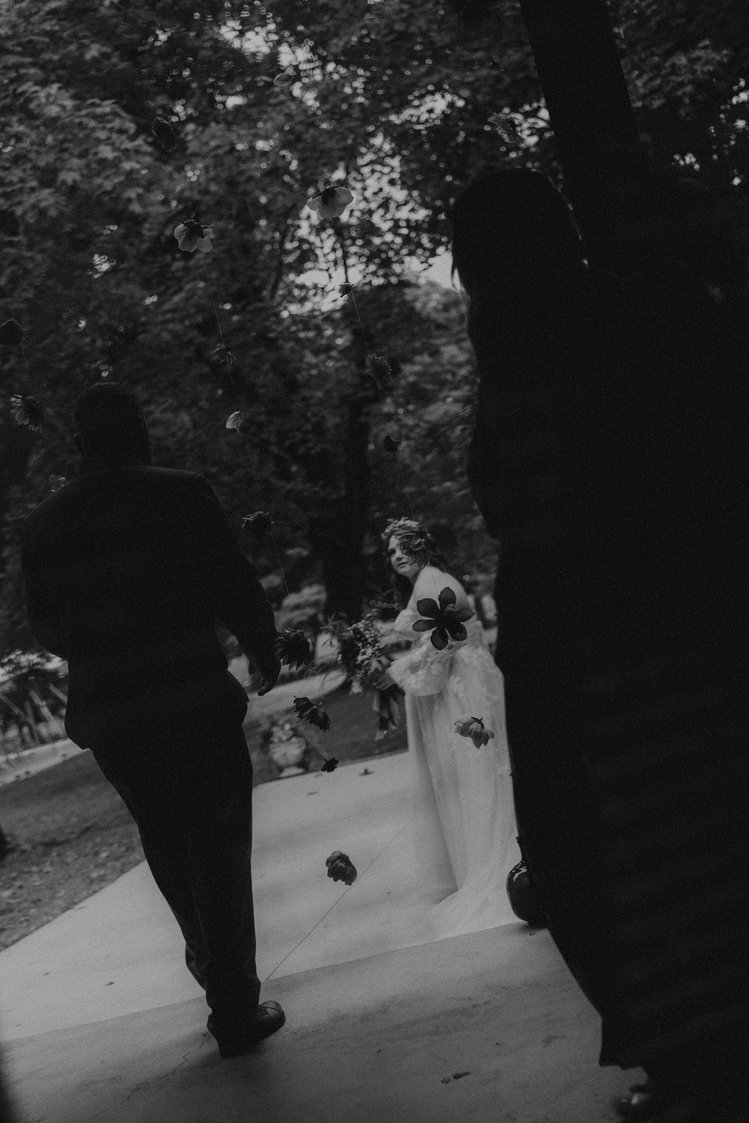 Black and white wedding photo of bride in white gown holding bouquet, groom walking toward her outdoors.
