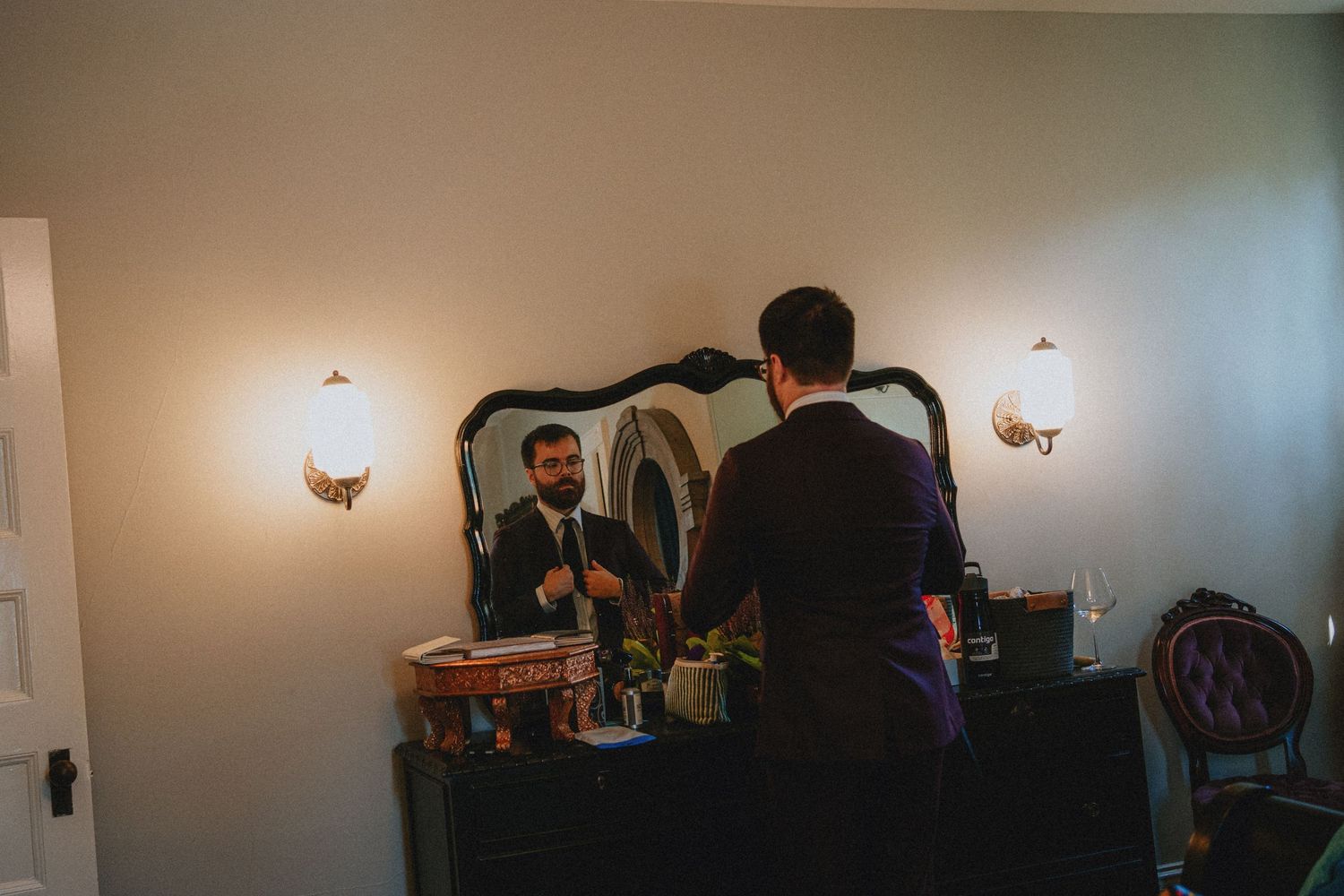 Groom adjusting tie in ornate mirror on dark dresser, flanked by wall sconces in elegant wedding preparation room.