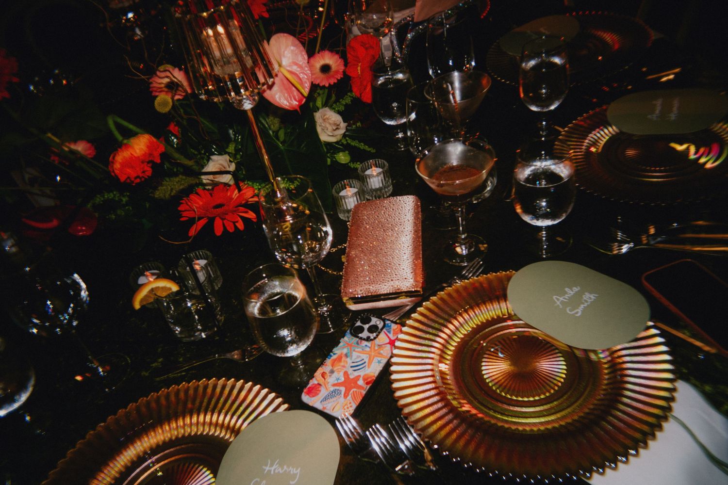 Elegant wedding table setting with gold charger plates, crystal glasses, and vibrant floral centerpiece in dark moody lighting.