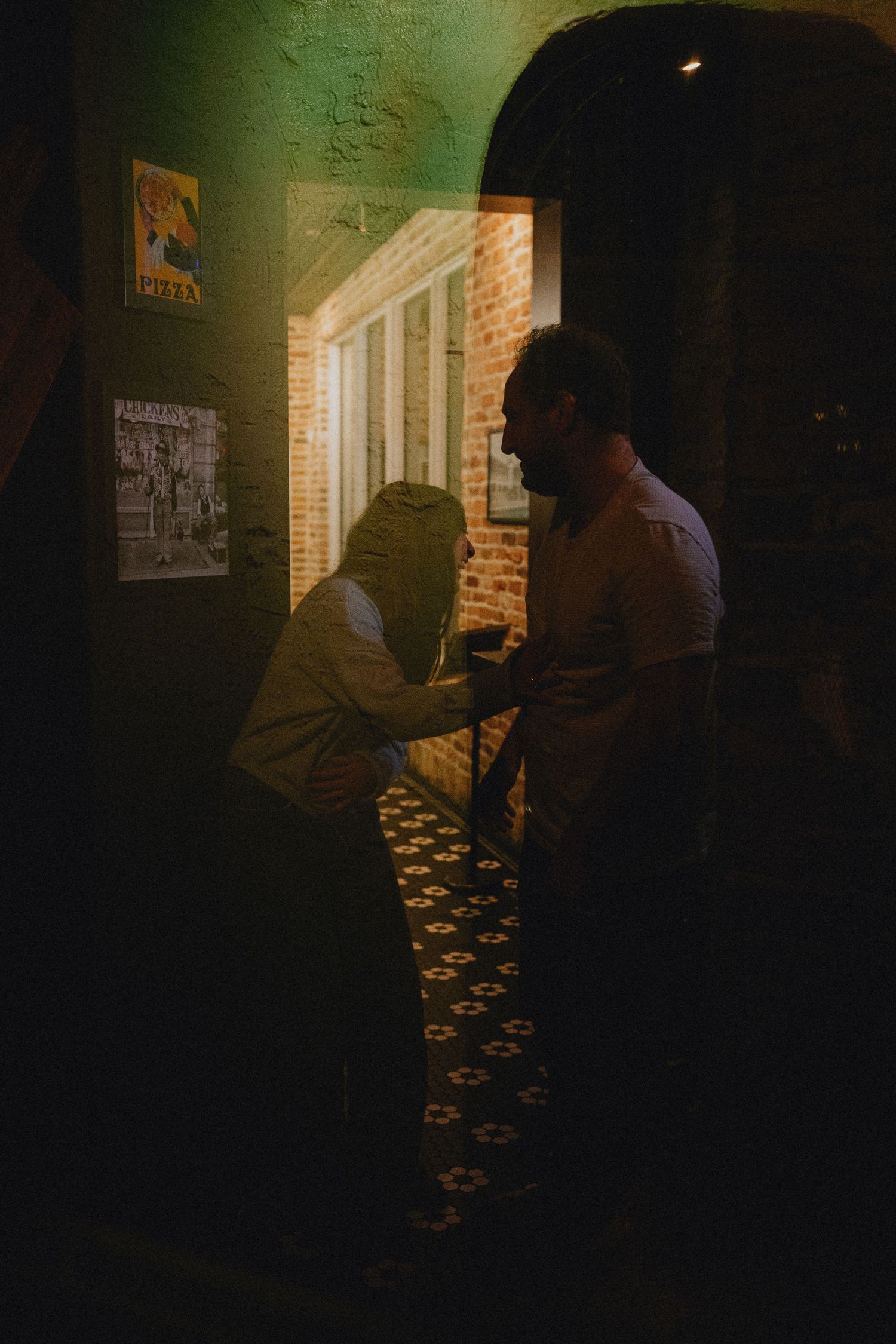 Two people share an intimate moment in a dimly lit corridor with brick walls and vintage posters.