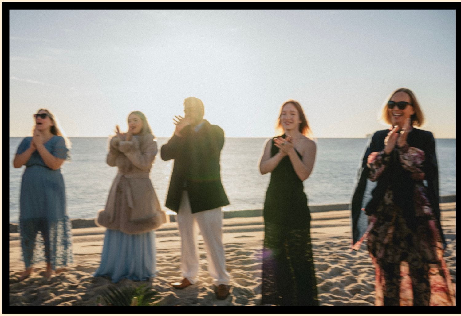 Group of elegantly dressed people clapping and celebrating on a sunny beach at golden hour.