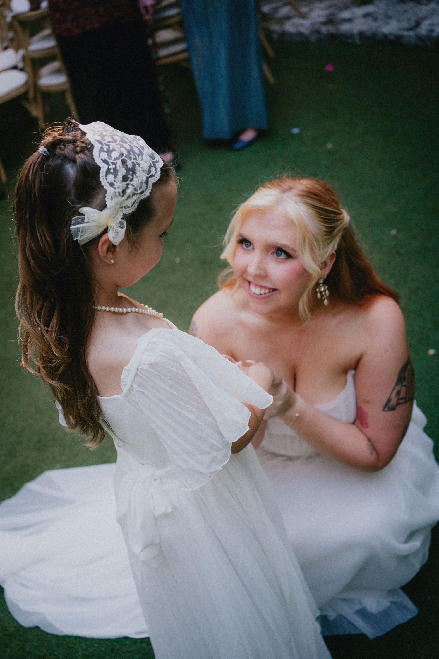 Two brides in white wedding gowns hold hands and smile at each other during their outdoor wedding ceremony.