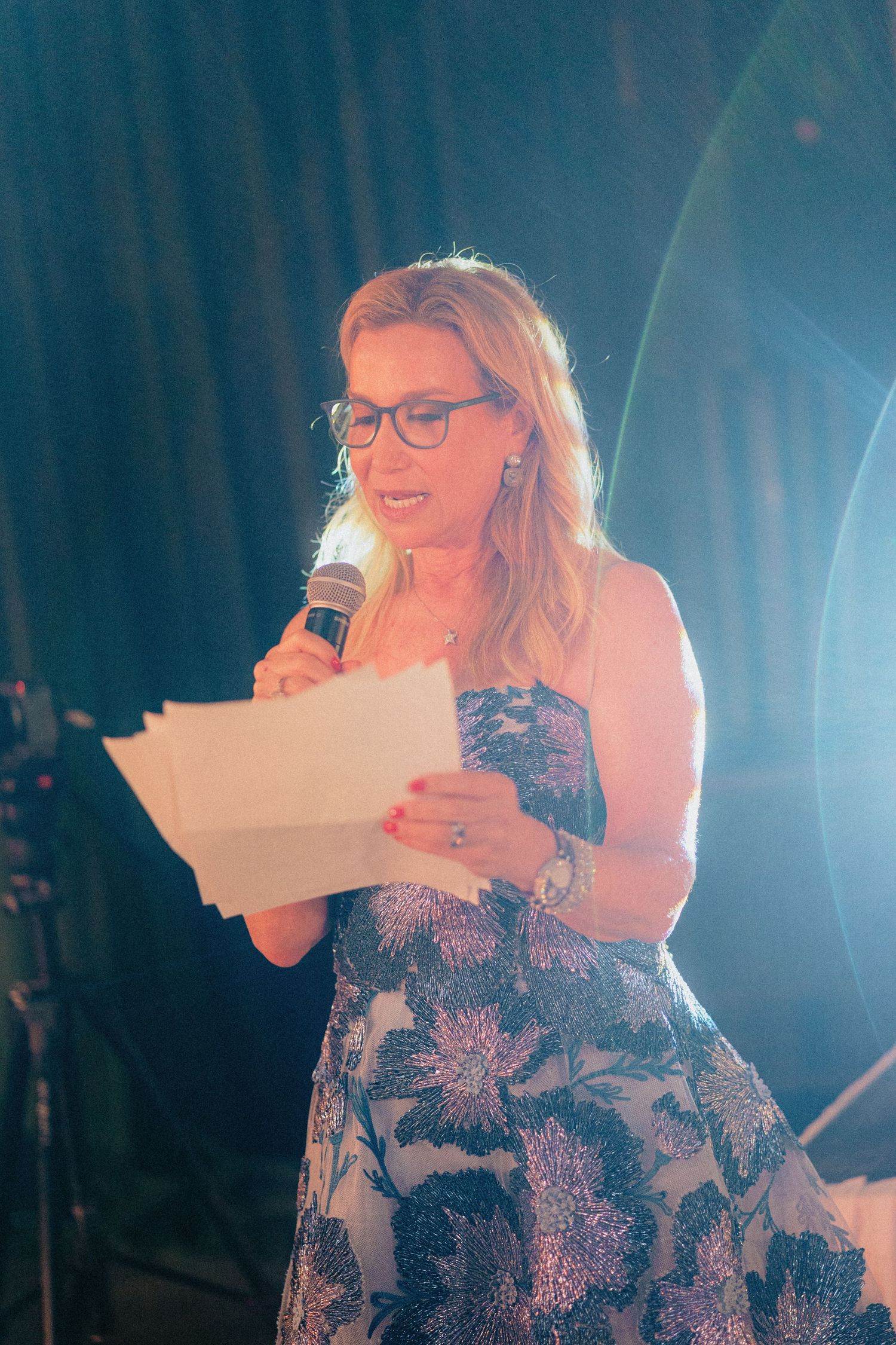 Woman in floral dress speaking at event, holding microphone and papers on stage with blue lighting.