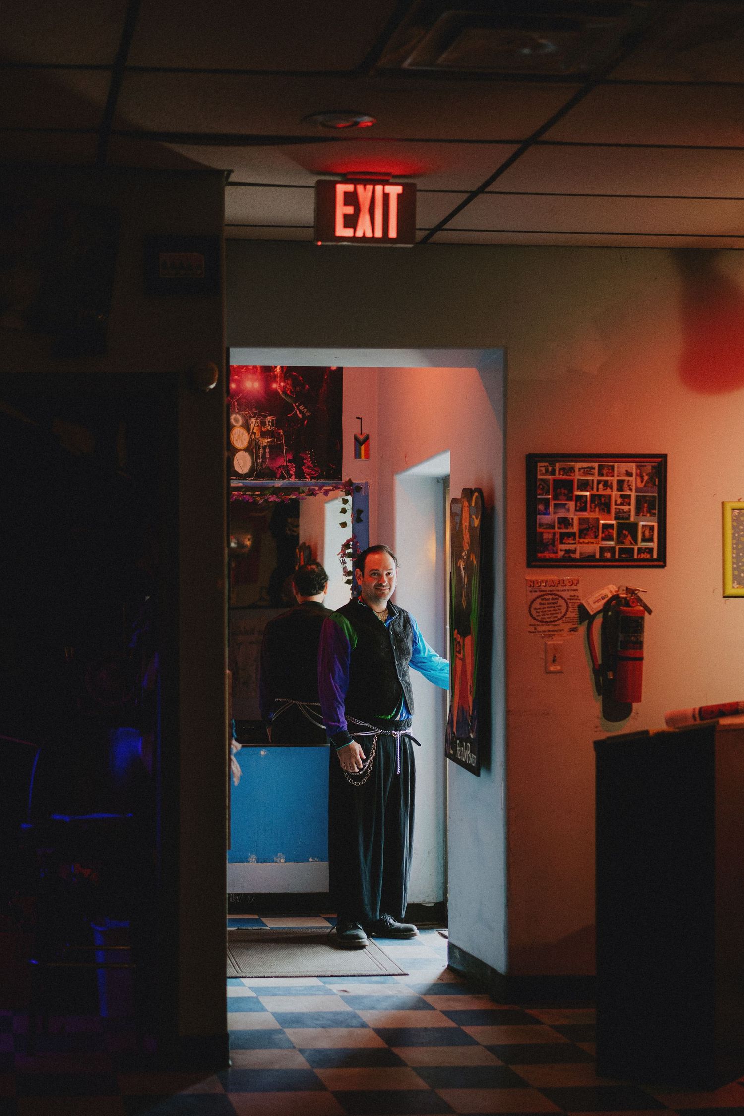 Man standing in a dimly lit hallway beneath a glowing red EXIT sign, with checkered floors and framed art on walls.