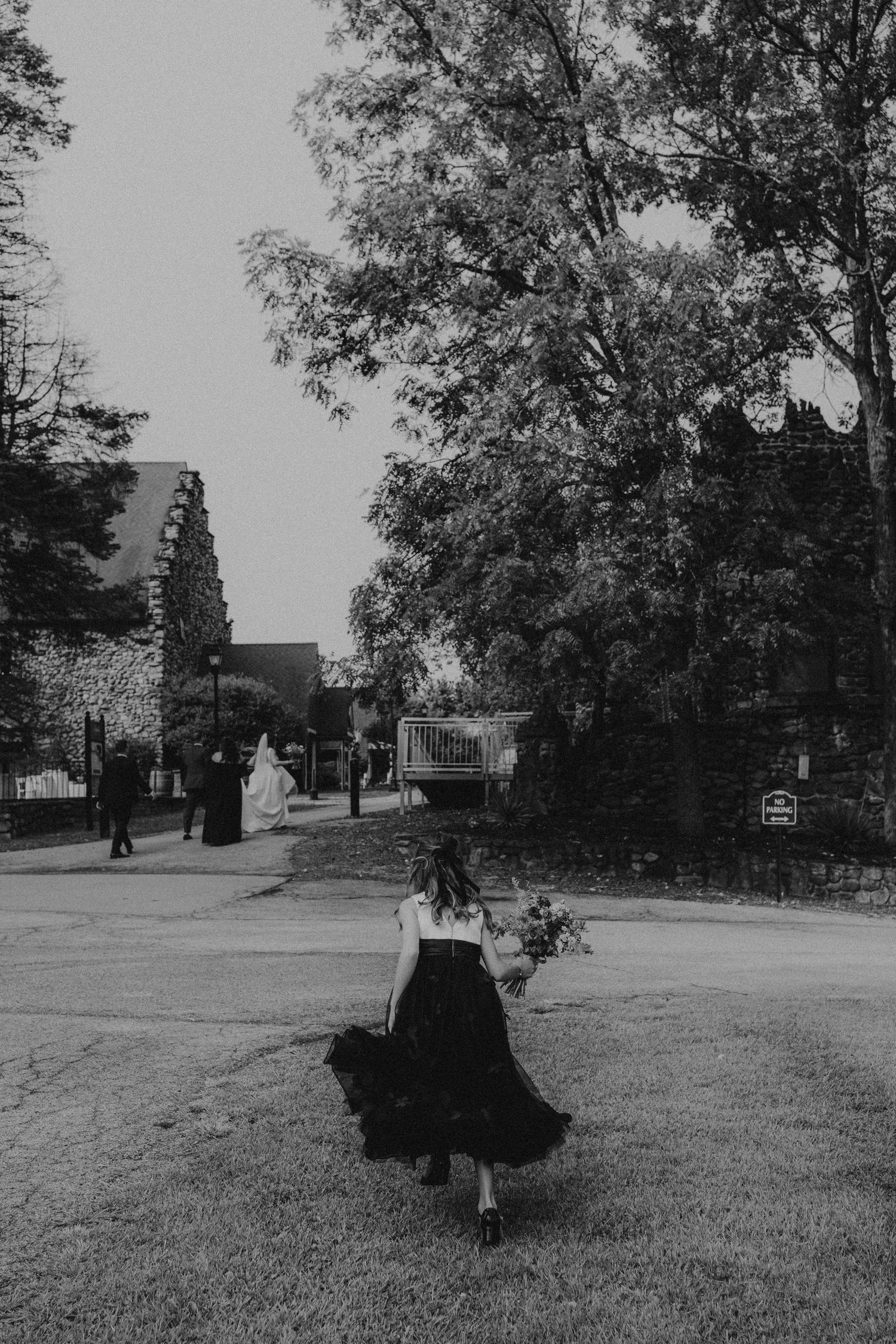 Black and white photo of a woman in a flowing skirt carrying flowers, walking toward a church on a tree-lined path.