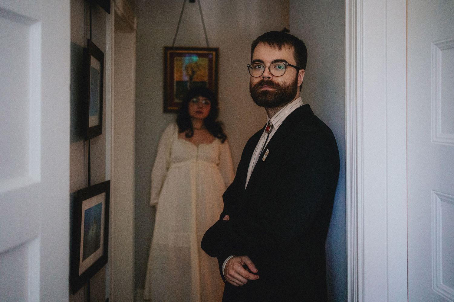 A bearded groom in a black suit stands in a hallway while a bride in a white dress stands behind him.