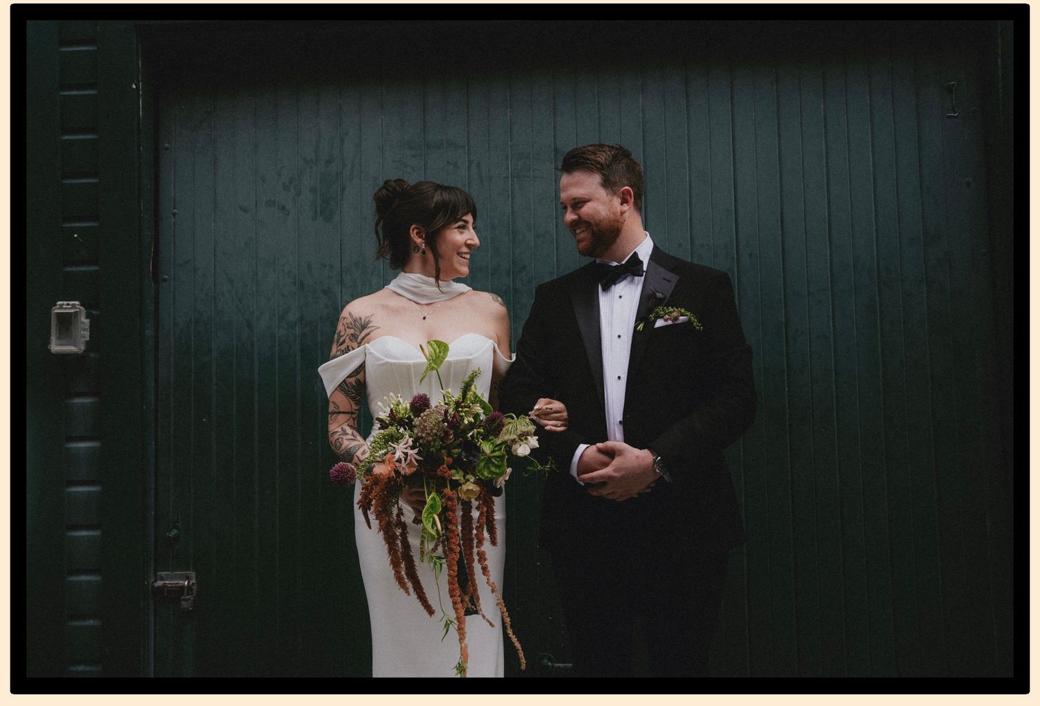 Bride and groom laughing together in front of a dark green door, bride holding a rustic bouquet.