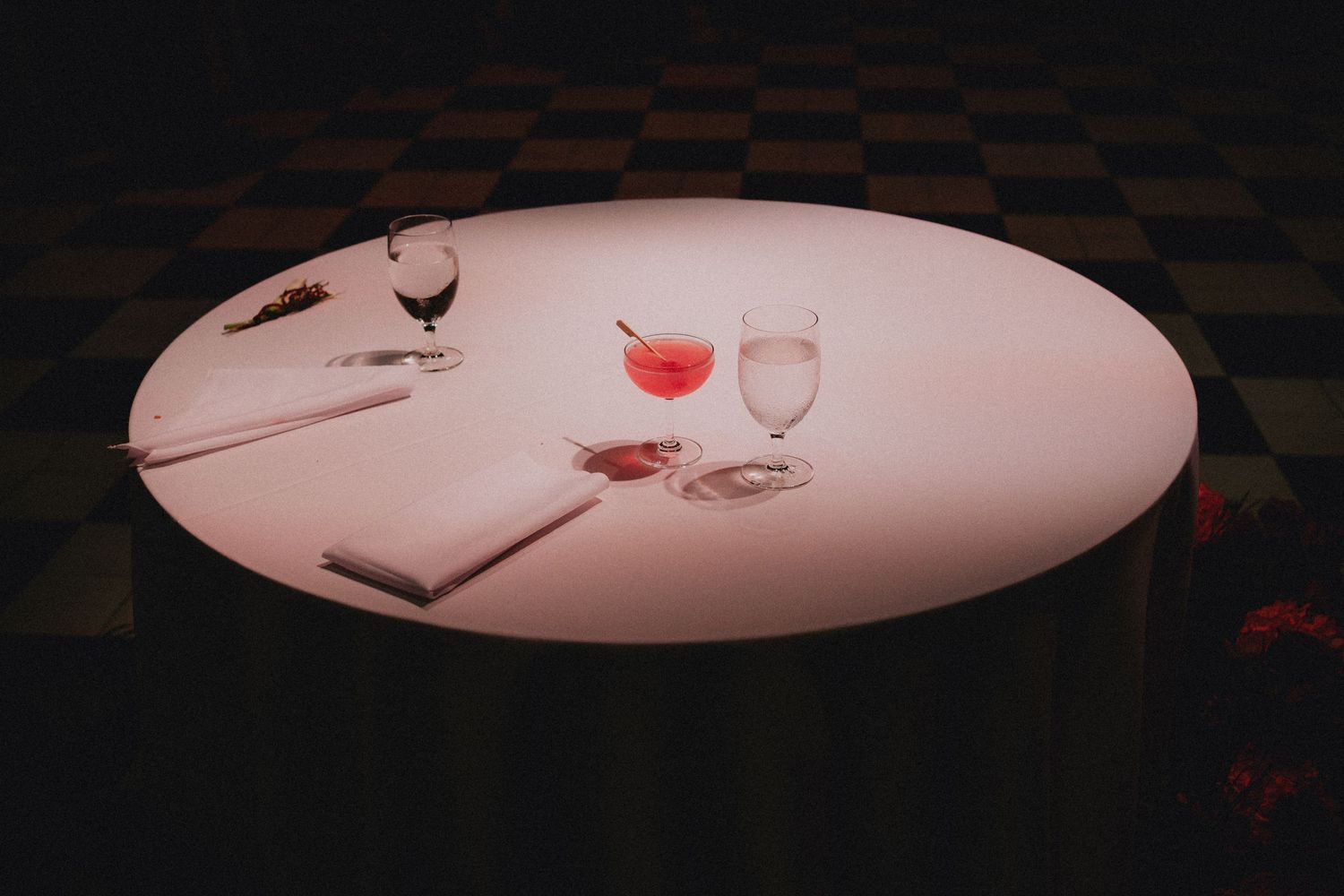 Elegant round white table with pink cocktail, wine glasses, and notepad in a dimly lit upscale restaurant.