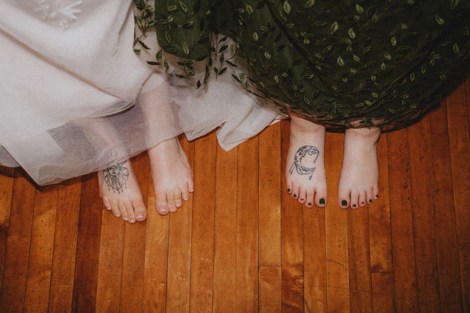 Two barefoot women in wedding attire stand on hardwood floor, one with a floral foot tattoo and the other with dark painted toenails.