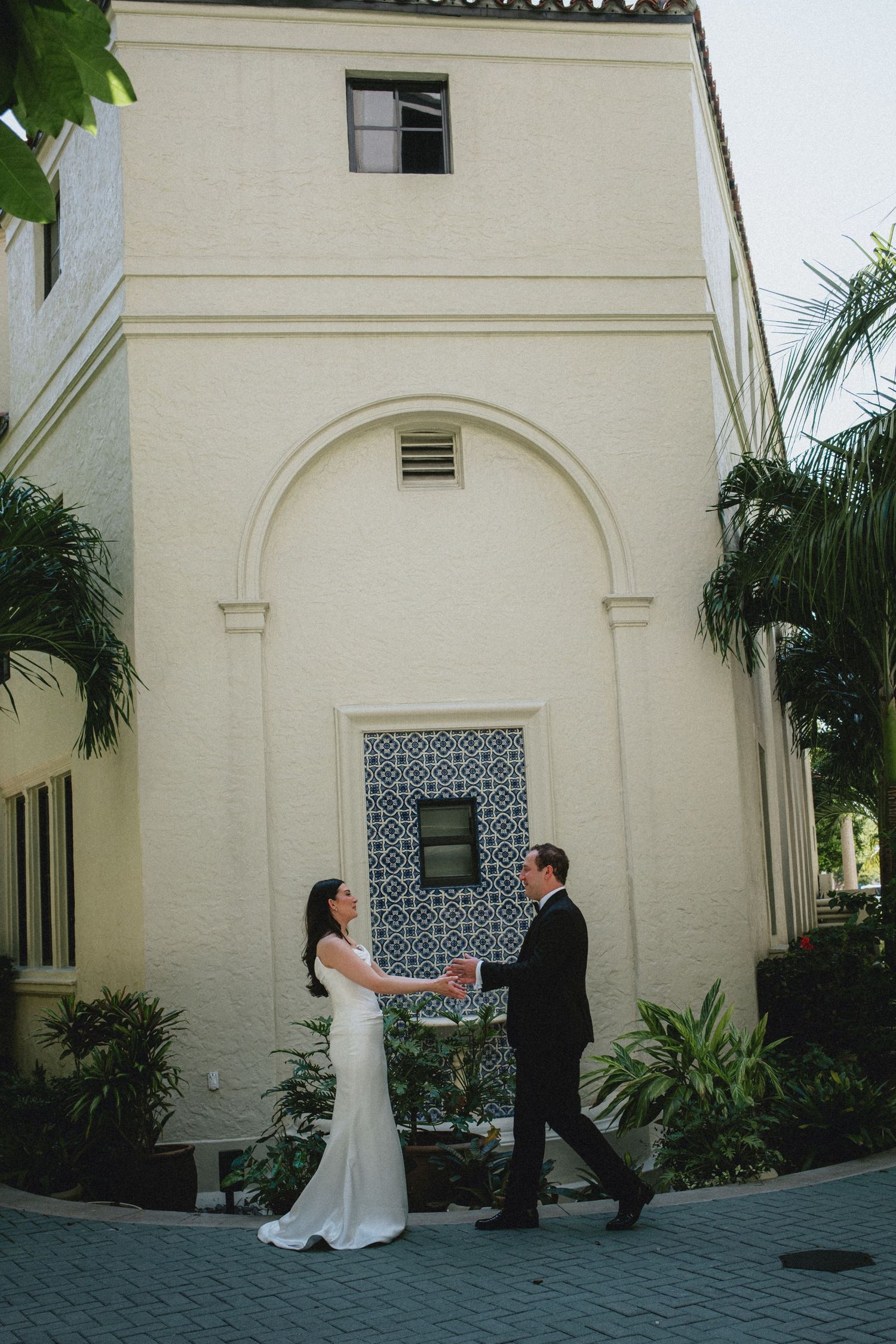 Bride and groom hold hands outside a Mediterranean-style building with blue tile door and tropical palms.