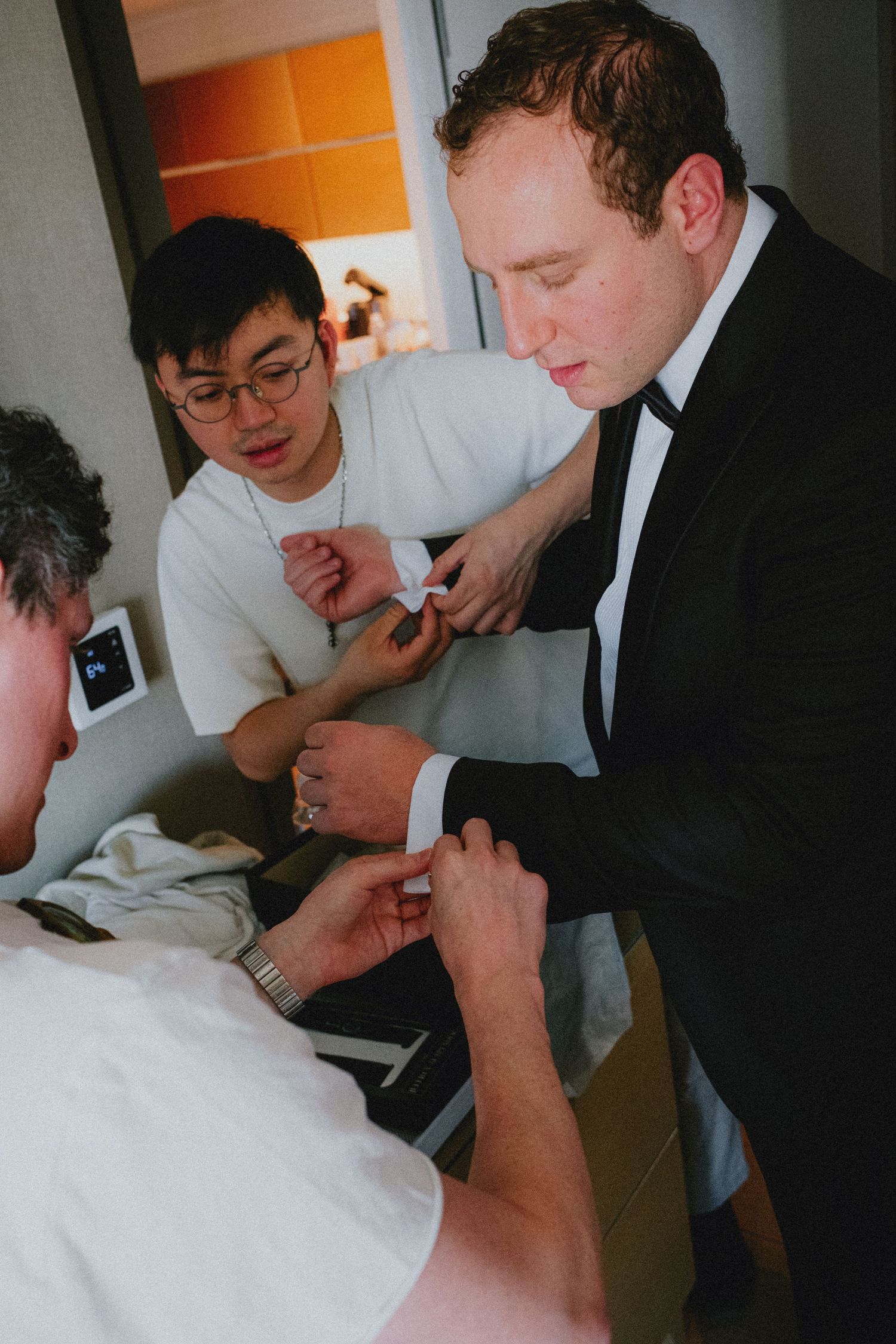 Groom in black tuxedo getting cufflinks fastened by groomsmen before wedding ceremony.