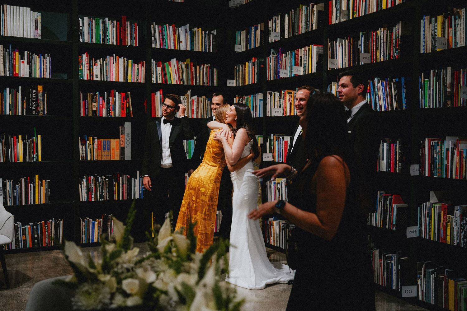 Couple shares a kiss during intimate library wedding ceremony surrounded by floor-to-ceiling bookshelves and guests.