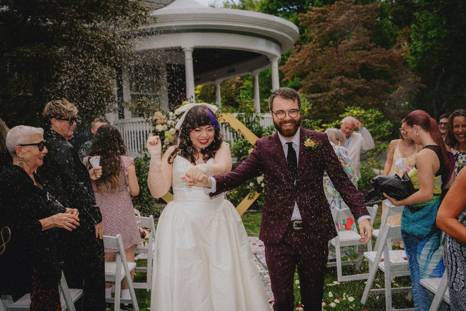 Bride and groom walk through confetti shower after outdoor wedding ceremony in garden with gazebo.