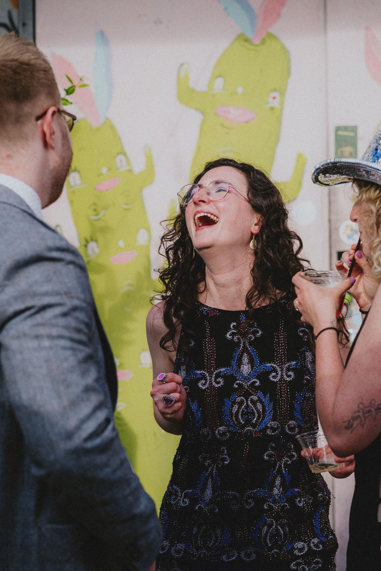 A woman in a blue embroidered dress laughs joyfully at a colorful event with friends nearby.