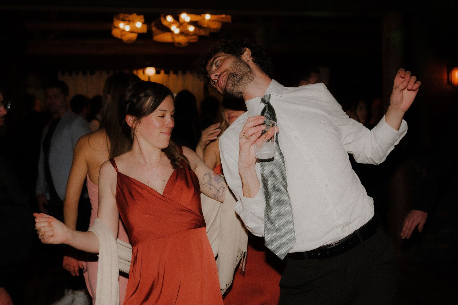 People dancing and moving in a dimly lit setting with someone wearing an orange dress visible in the scene.