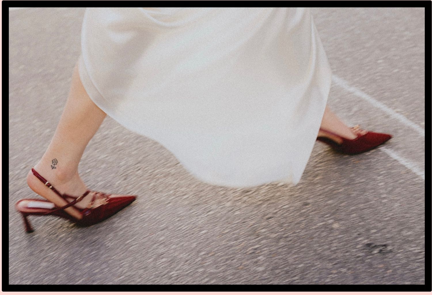 Woman walking in red strappy heels with ankle tattoo, wearing a flowing white dress on pavement.