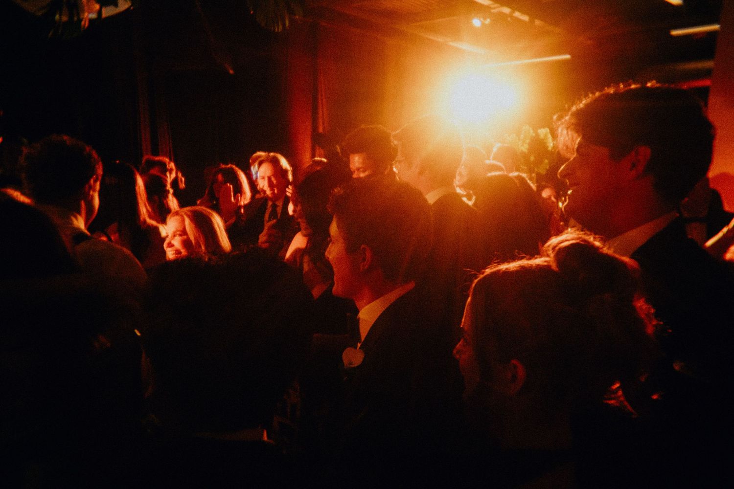 Crowd of people mingling at a dimly lit evening event with warm orange lighting in the background.
