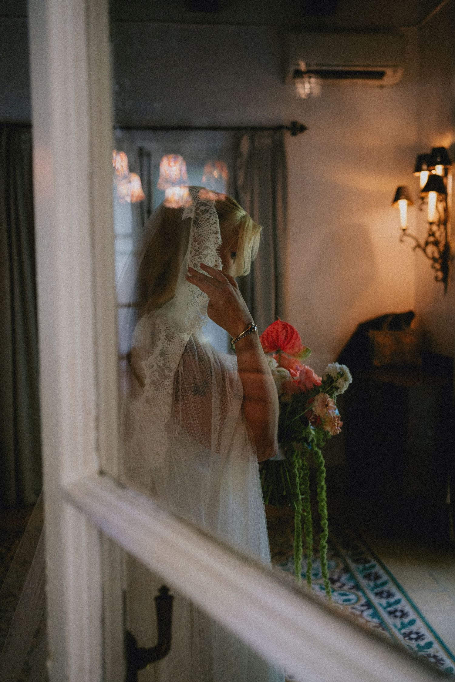 Bride in lace wedding dress and veil holding a pink floral bouquet, reflected in a window with warm ambient lighting.
