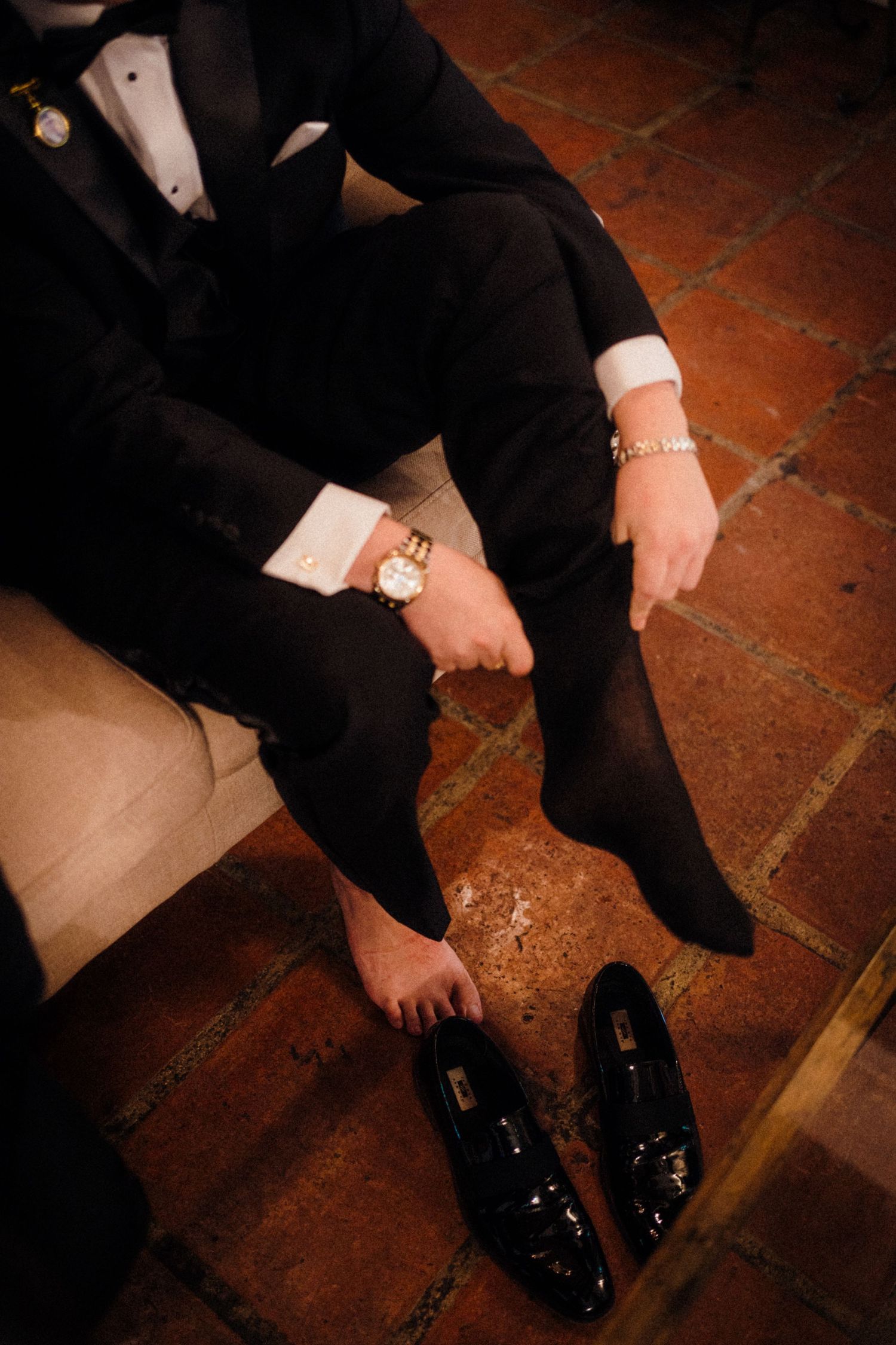 A groom in a black tuxedo puts on dress socks beside patent leather loafers on a terracotta tile floor.