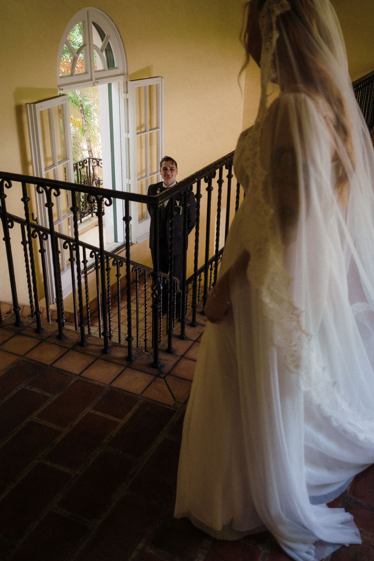 Bride in lace wedding gown and veil watches groom descend ornate staircase in elegant venue.