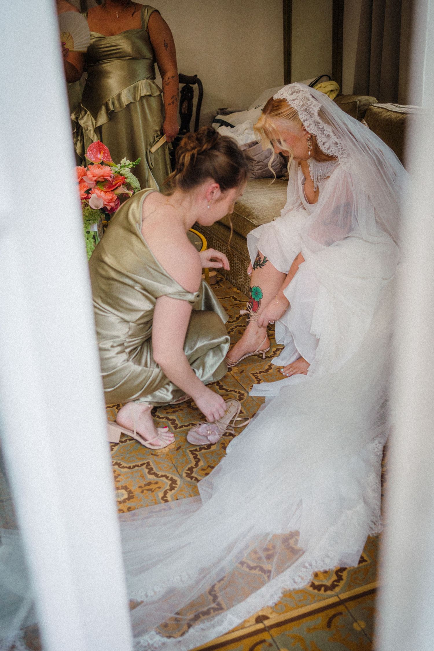 Bridesmaid in gold dress helping bride put on shoes before wedding ceremony.