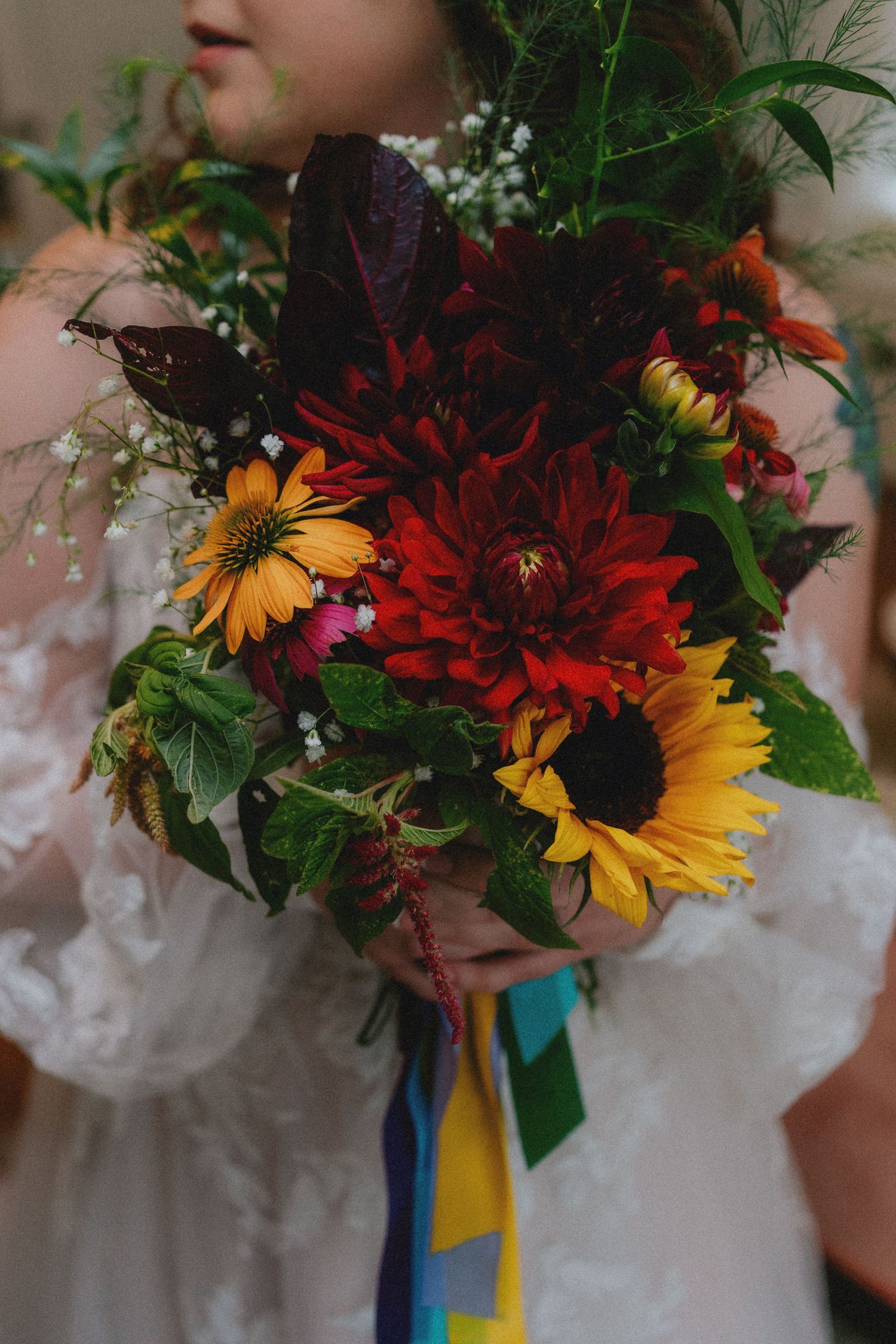 Bride holding a vibrant wedding bouquet with sunflowers, red dahlias, and greenery, tied with a colorful ribbon.