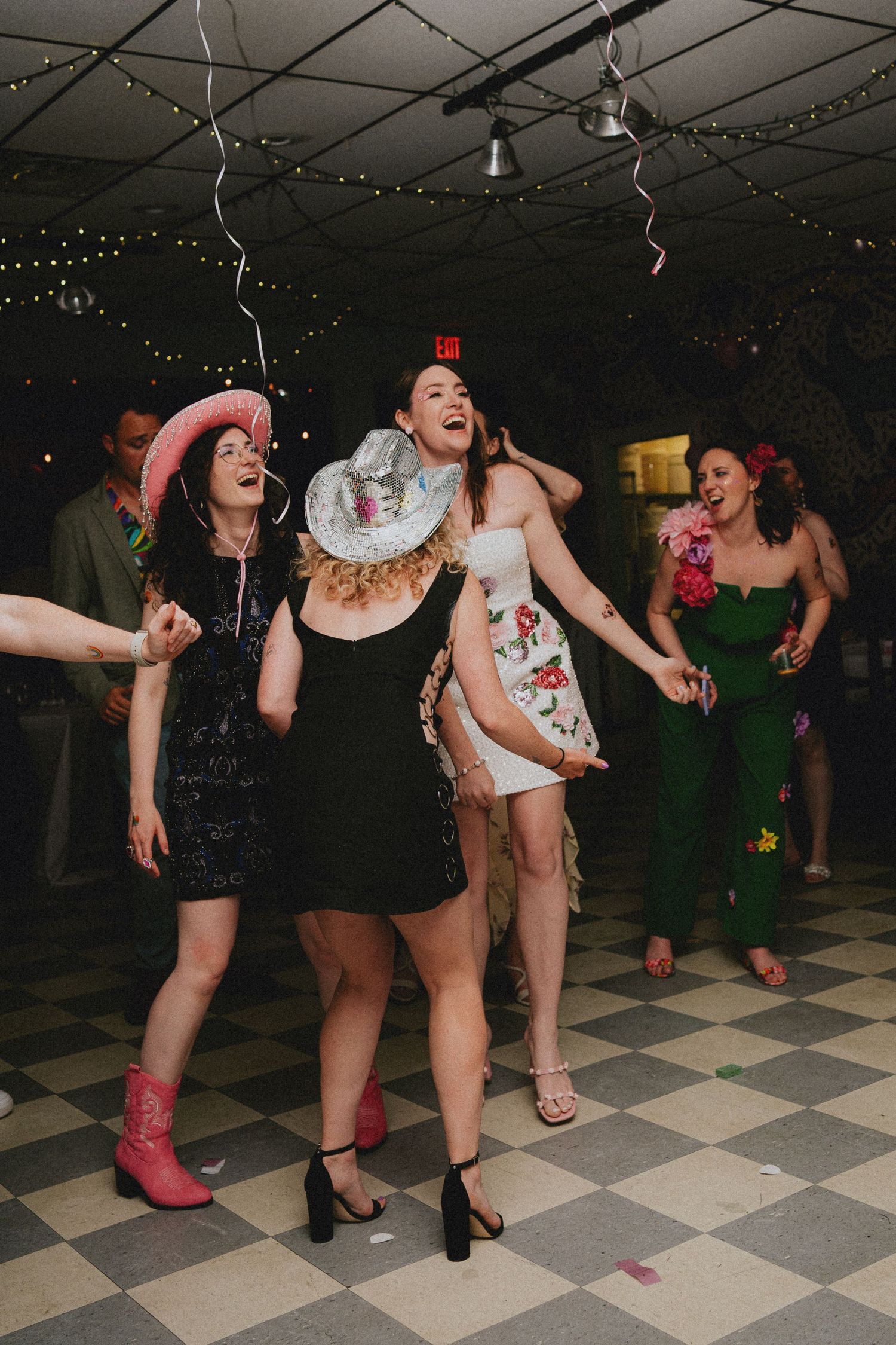 Women dancing joyfully at a party on checkered floor, wearing festive hats and colorful dresses under string lights.