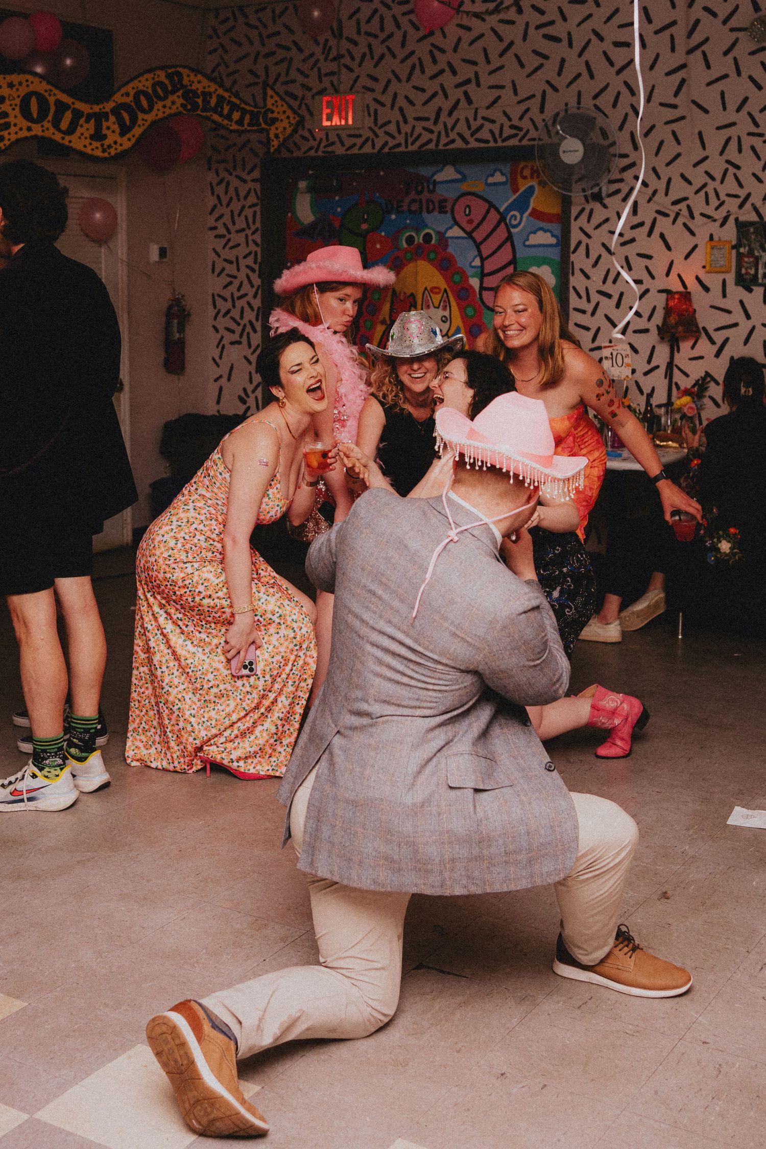 People in pink hats and festive outfits dancing at a colorful party venue with confetti.