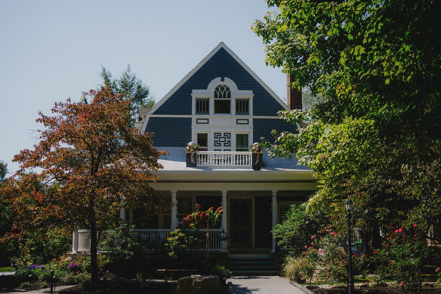 Blue Victorian house with white trim, wraparound porch, and lush surrounding trees on a sunny day.