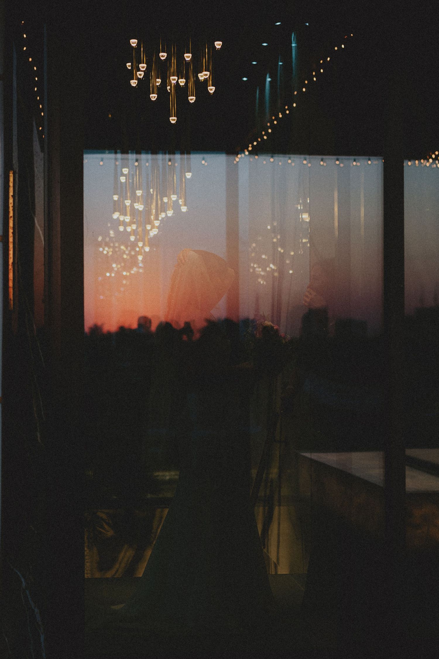 Moody interior view through sheer curtains revealing a stunning orange sunset skyline with warm string lights above.