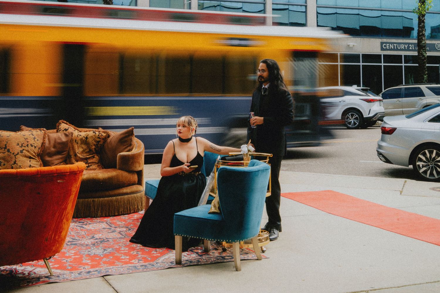Two elegantly dressed people pose outdoors with vintage furniture as a yellow bus blurs past on a city street.