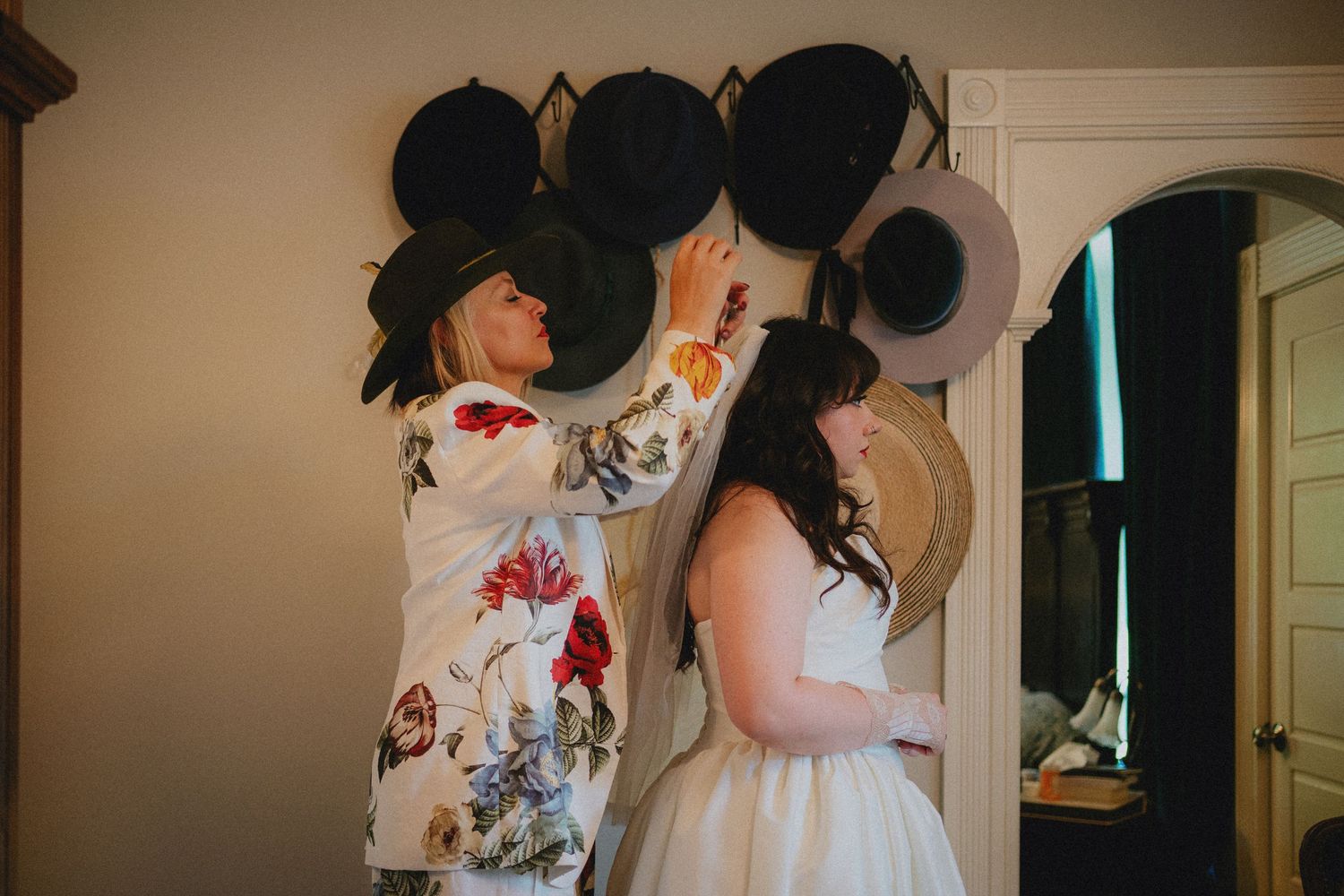 A bride in a white dress tries on hats with a woman in a floral jacket in a rustic room with hats displayed on the wall.