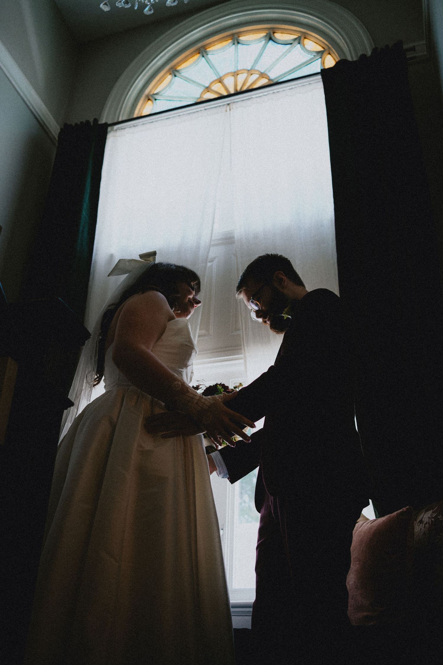 Silhouetted bride and groom exchanging rings during wedding ceremony before large arched window with sheer curtains.