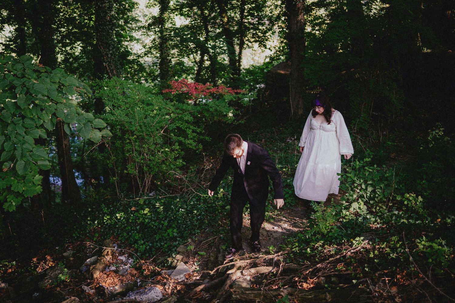 Bride in white gown and groom in black suit walking through lush green forest on their wedding day.
