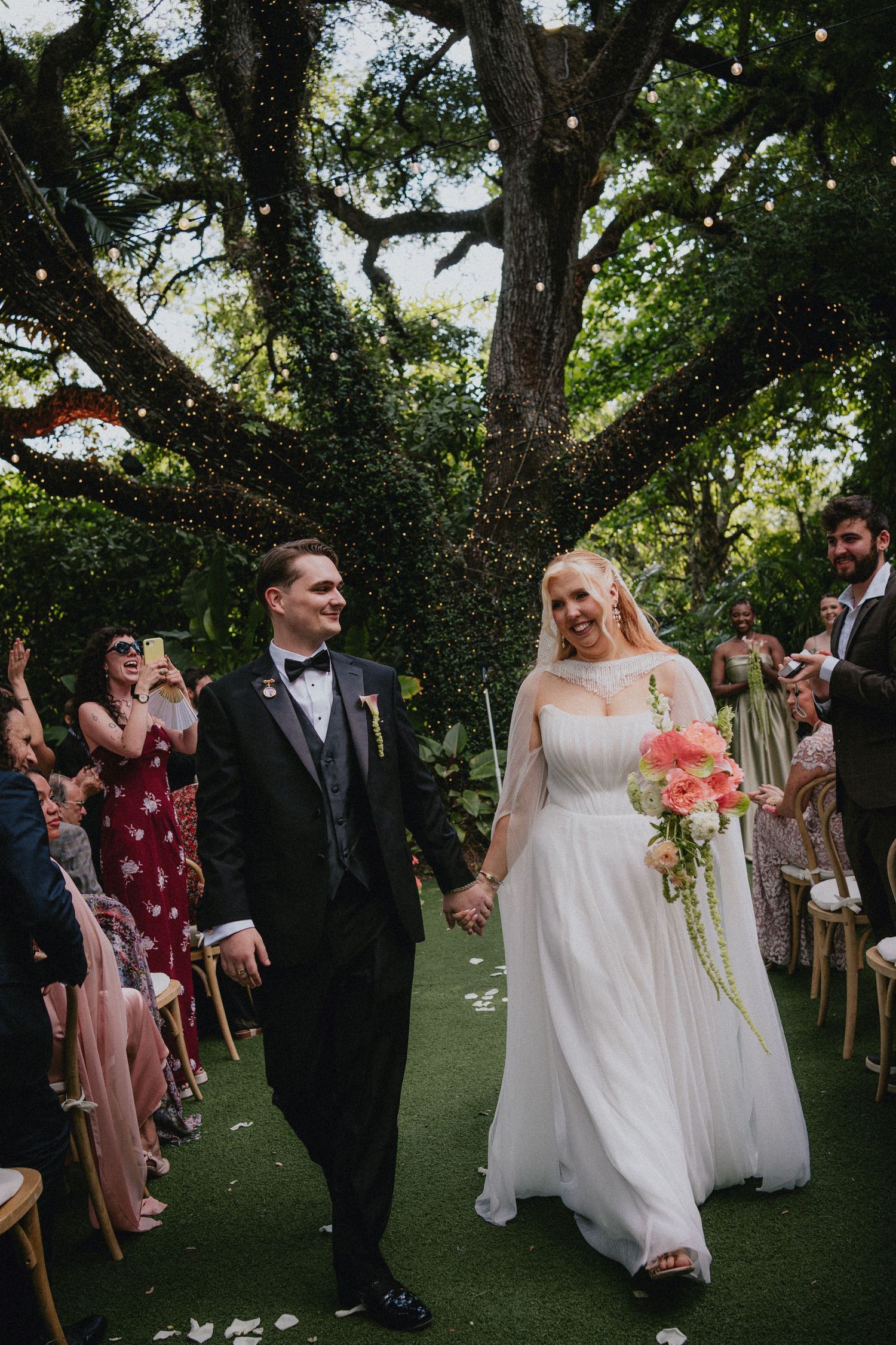 Bride and groom walk hand-in-hand down outdoor aisle under oak tree with string lights, guests cheering around them.