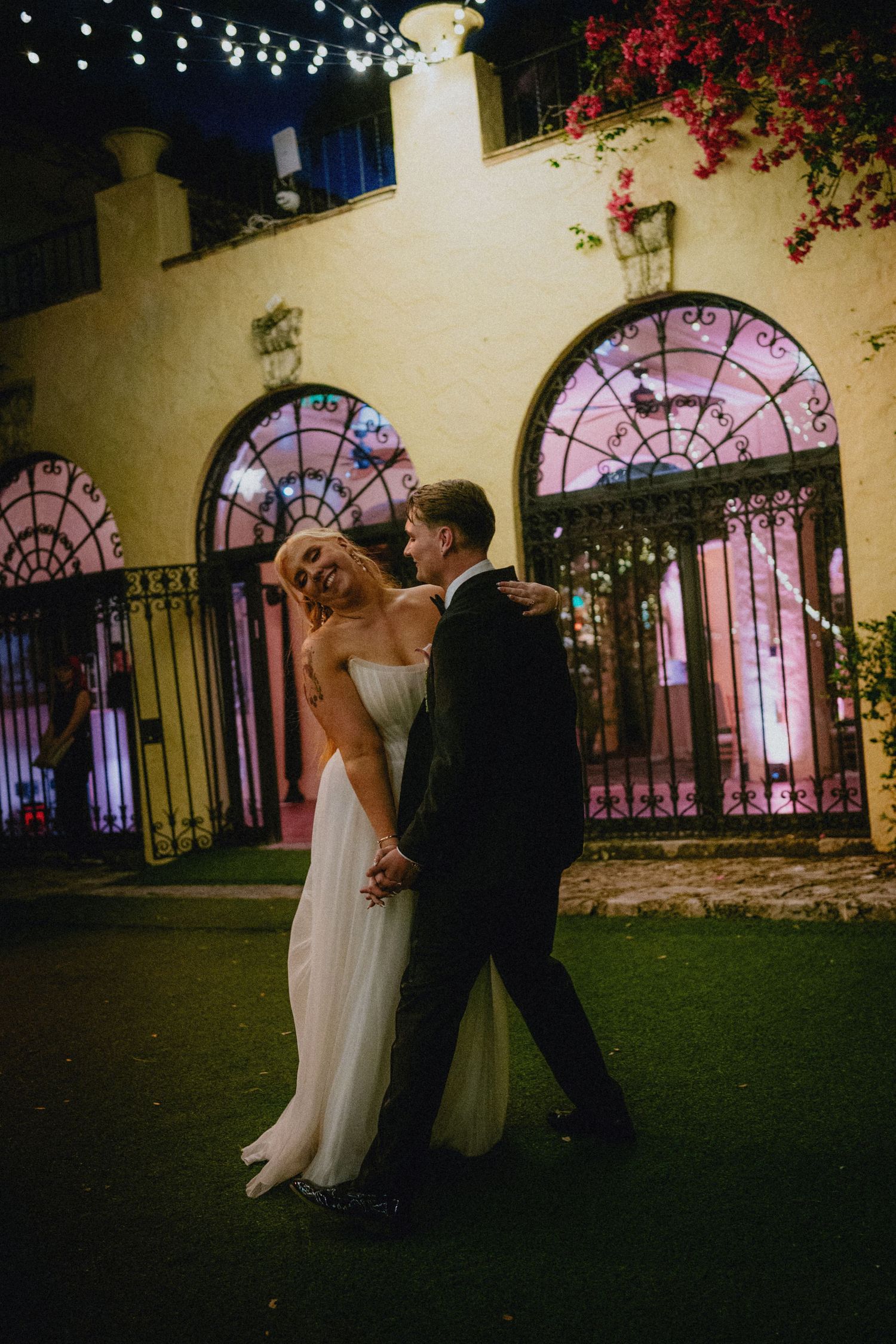Bride and groom share a romantic dance outside an elegant venue with arched doors and string lights at night.
