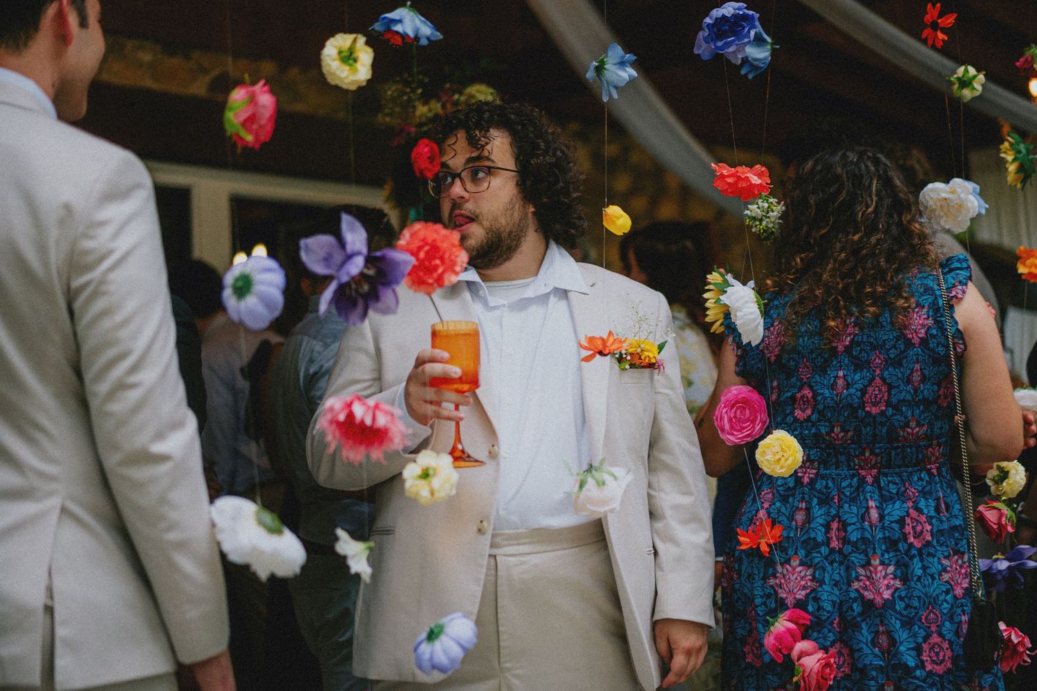 Man in beige suit holding cocktail at colorful floral wedding reception with hanging paper flower decorations.