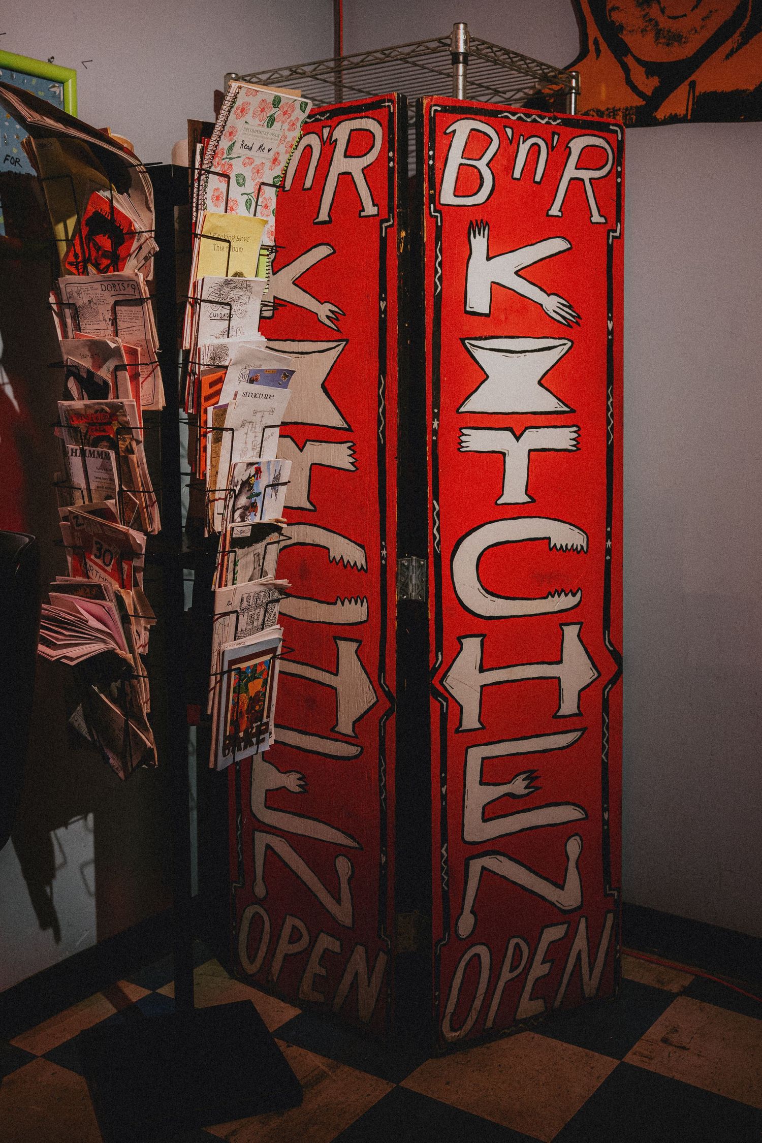 Red folding sign for B n R Kitchen displaying OPEN on checkered floor near magazine rack.
