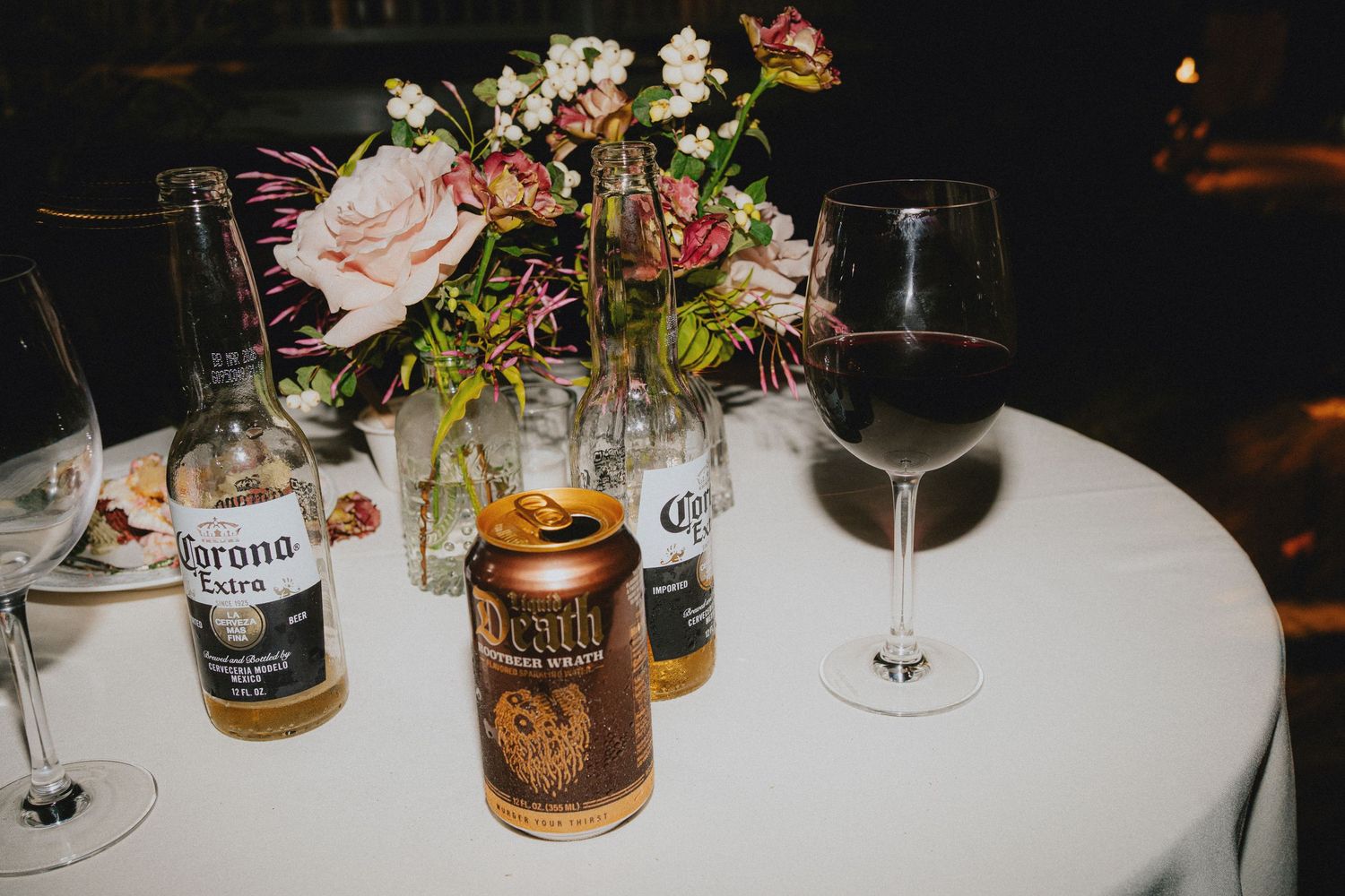 Corona Extra beer bottles, a Tecate can, and a glass of red wine on a white tablecloth with floral centerpiece.