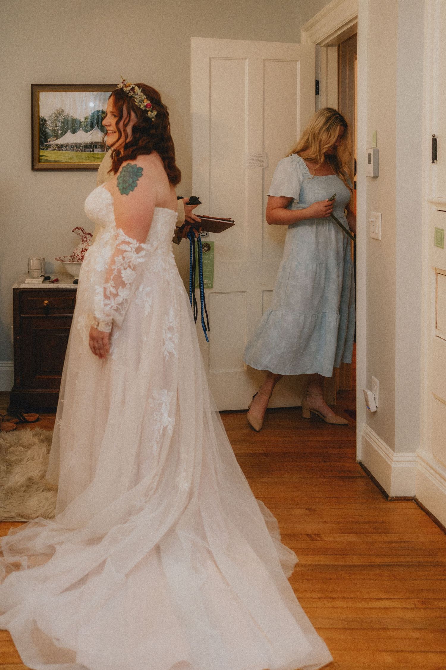 Bride in lace wedding gown with floral crown stands in room while bridesmaid in blue dress looks on.