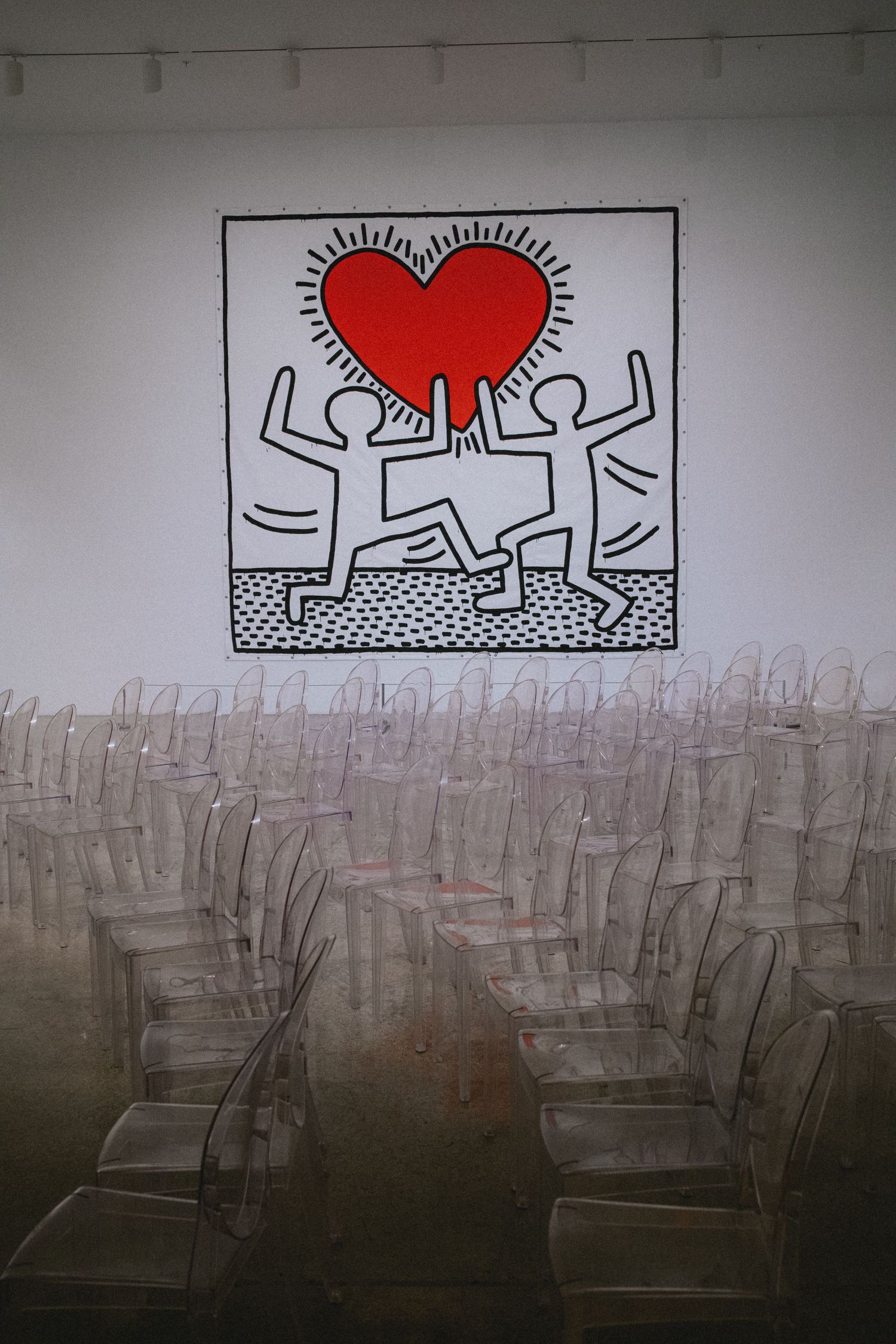 Keith Haring-style mural of two figures holding a red heart on white wall above rows of clear acrylic chairs.