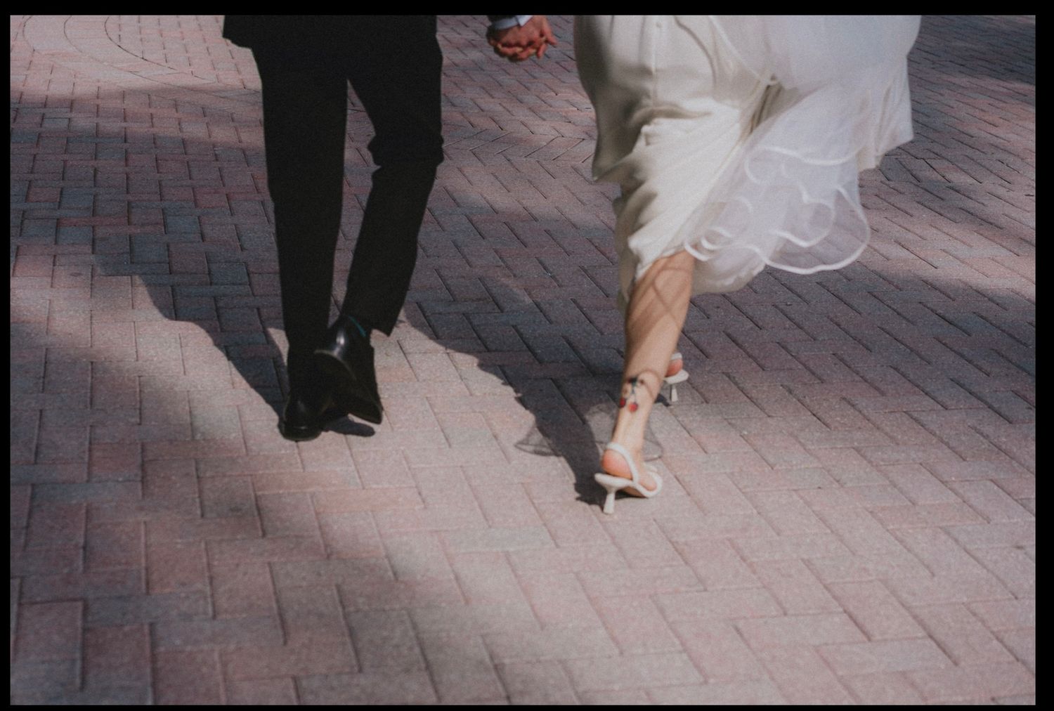 Close-up of a bride in white dress and heels walking alongside a groom in black suit on a brick pathway.