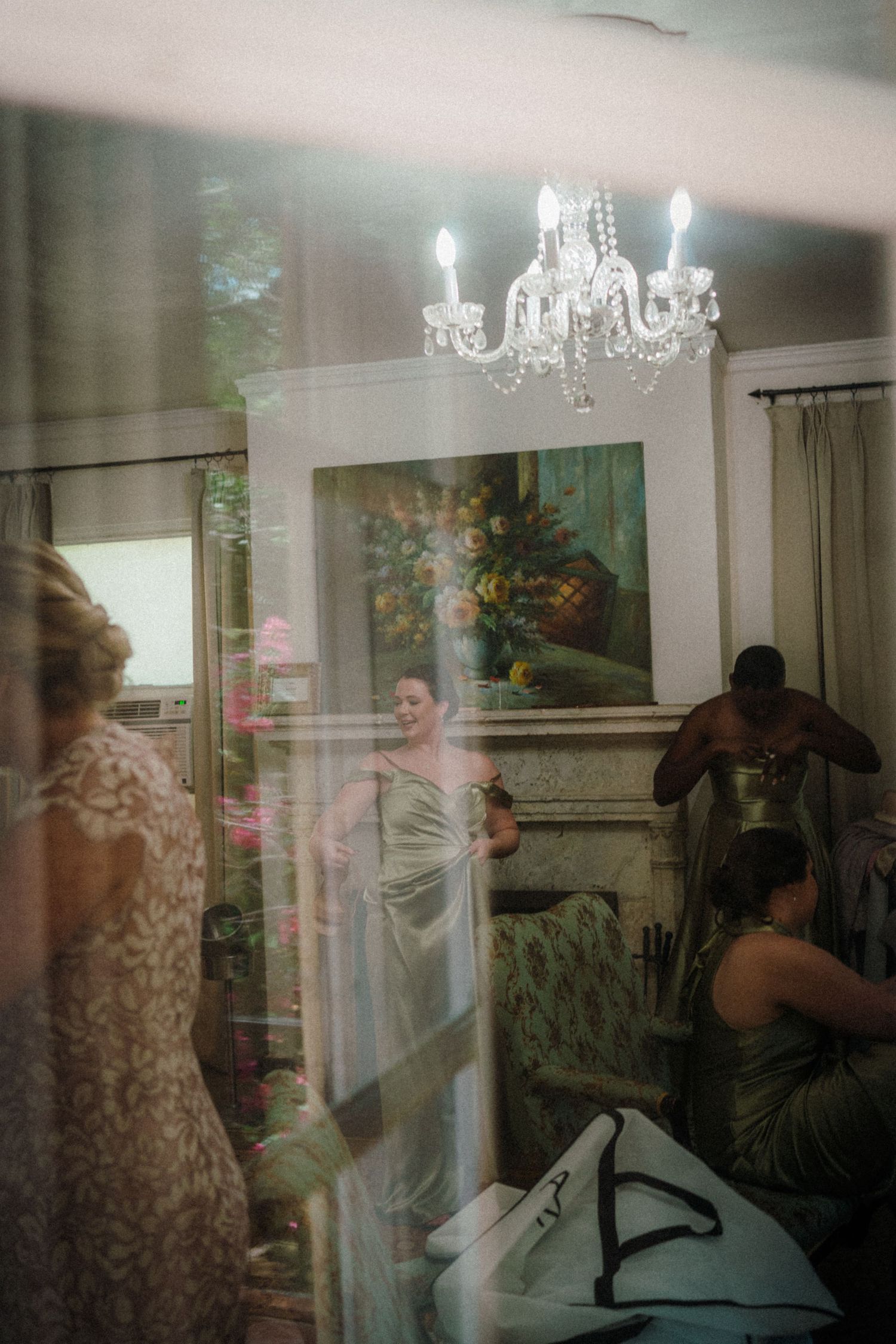 Bridesmaids helping bride get ready in elegant room with crystal chandelier and floral painting.