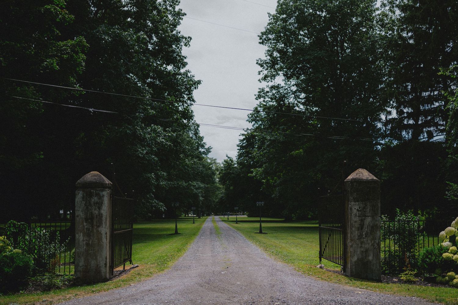 Stone gate pillars flank a tree-lined gravel driveway leading to a lush green estate on an overcast day.