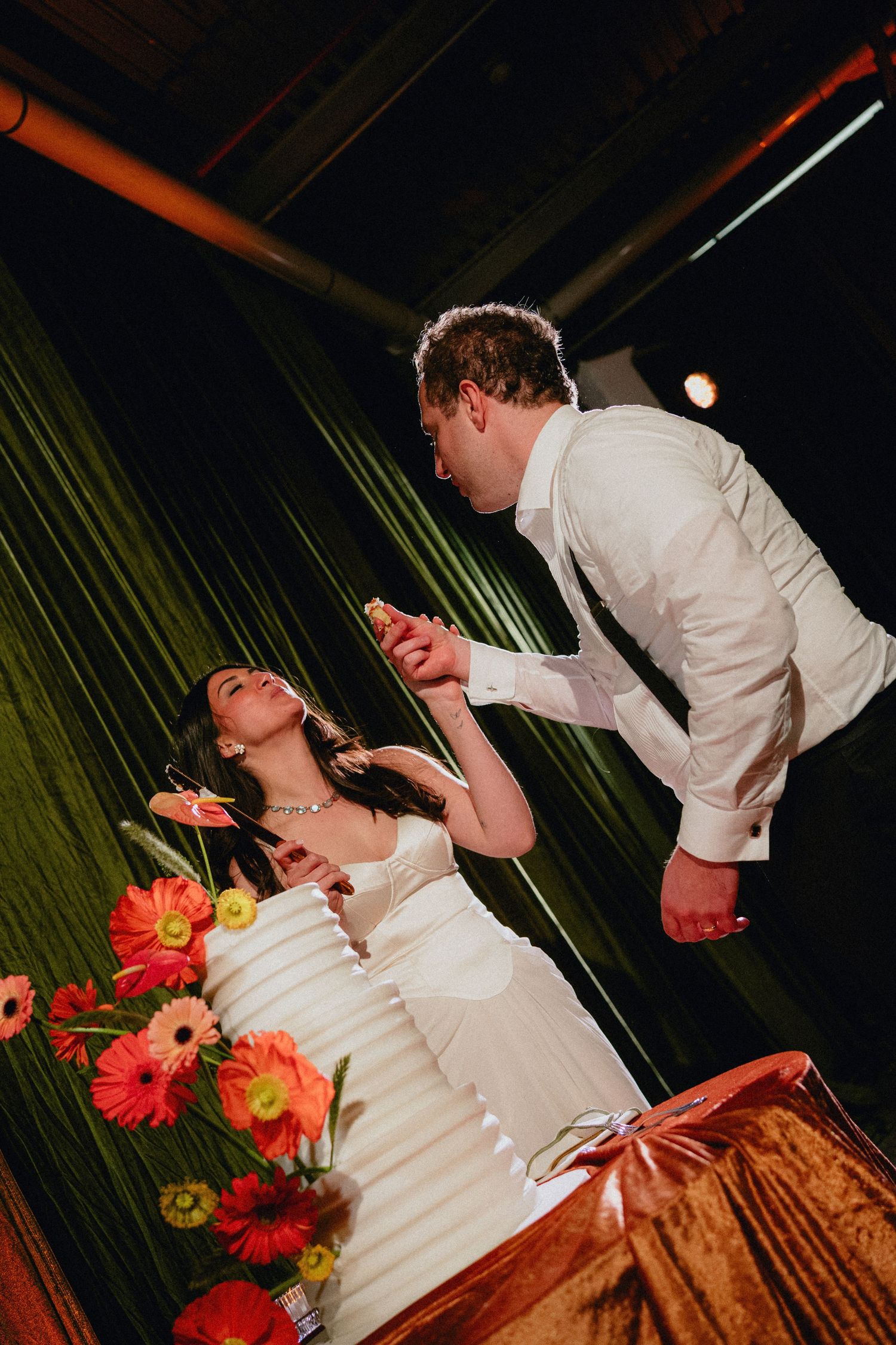 Bride and groom cutting their wedding cake adorned with colorful gerbera daisies at a romantic indoor reception.