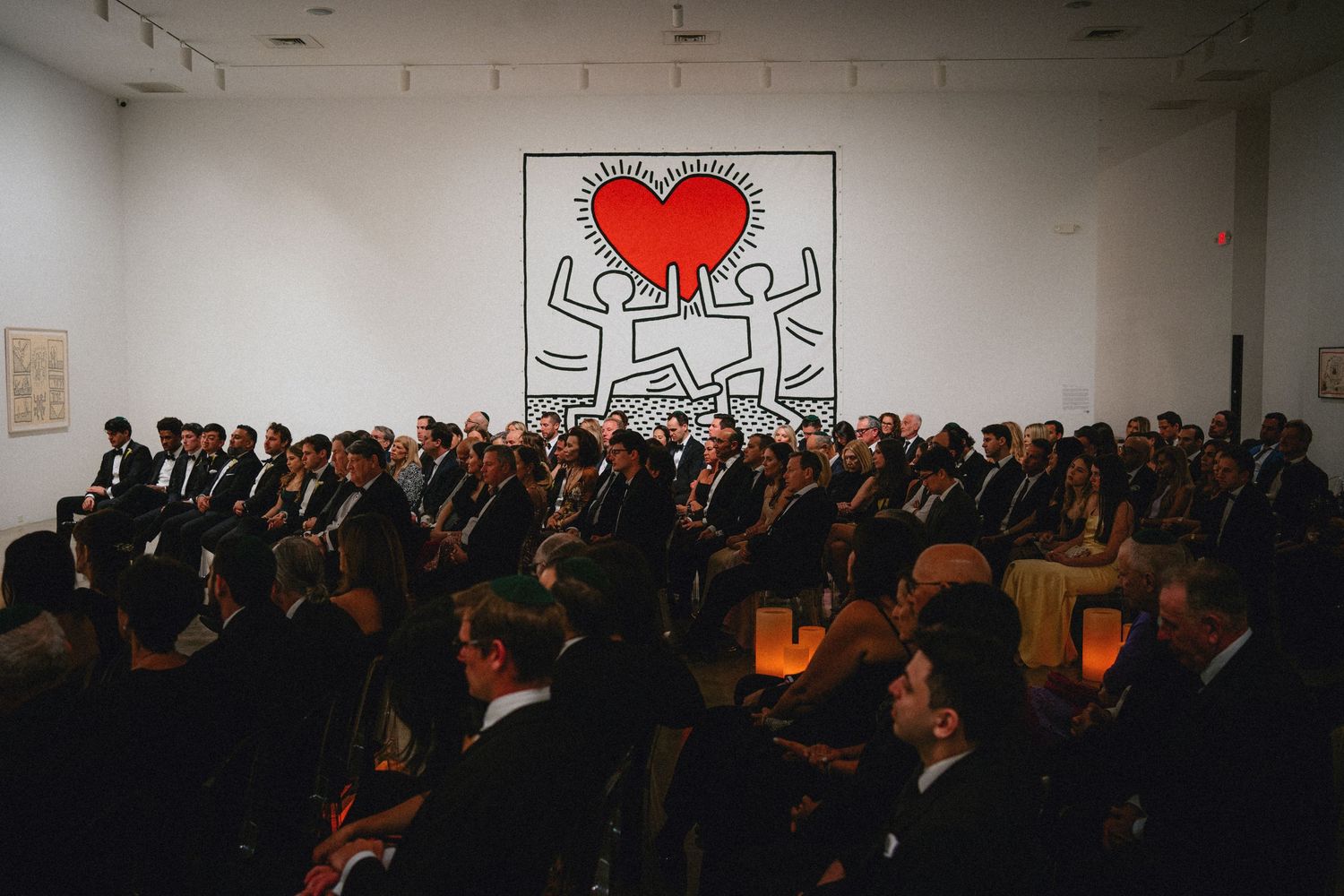Formal event crowd seated before a large Keith Haring heart artwork on a white gallery wall with candlelit tables.