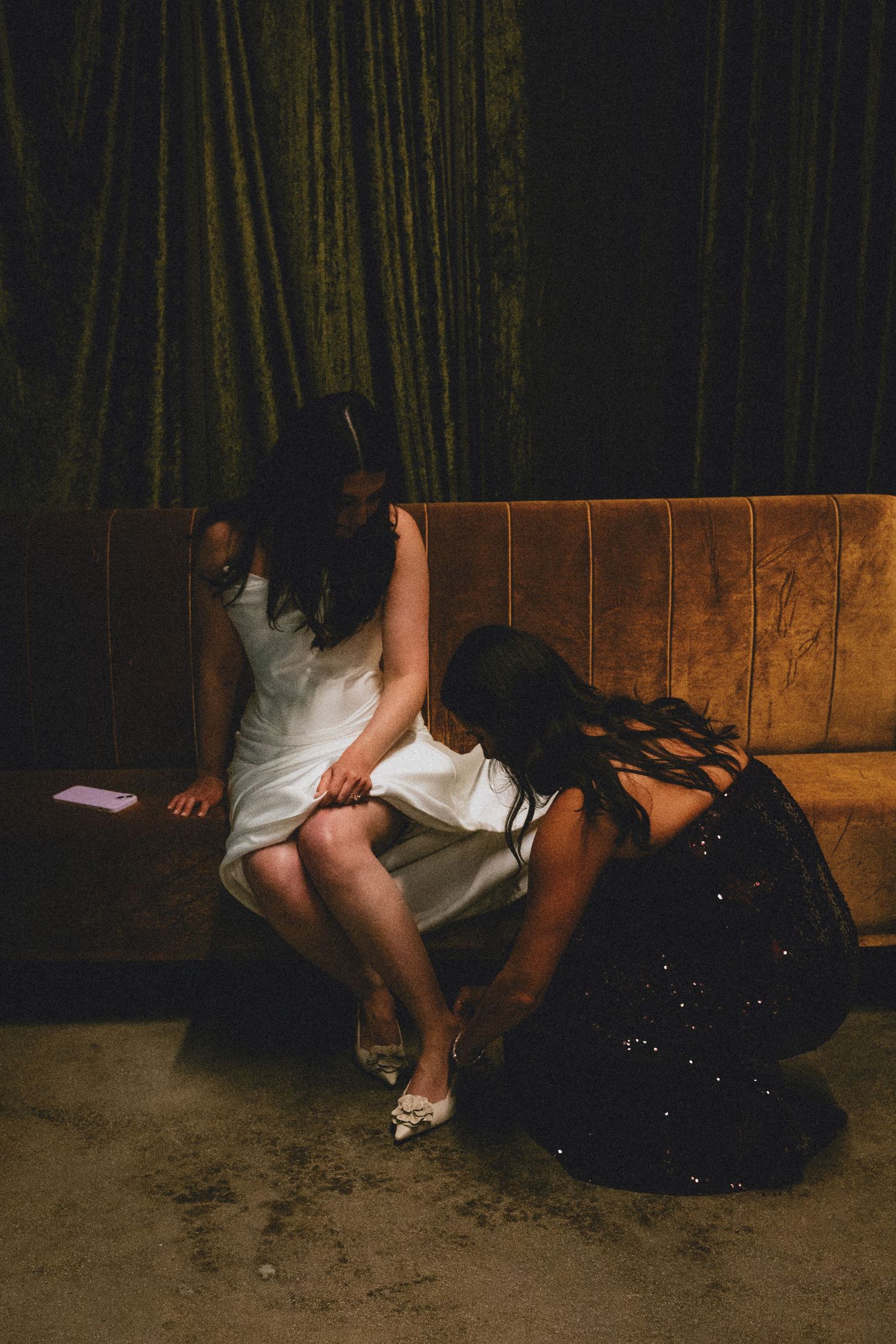 A woman in a white dress sits on a velvet booth while another in a black gown adjusts her shoe in a dimly lit venue.