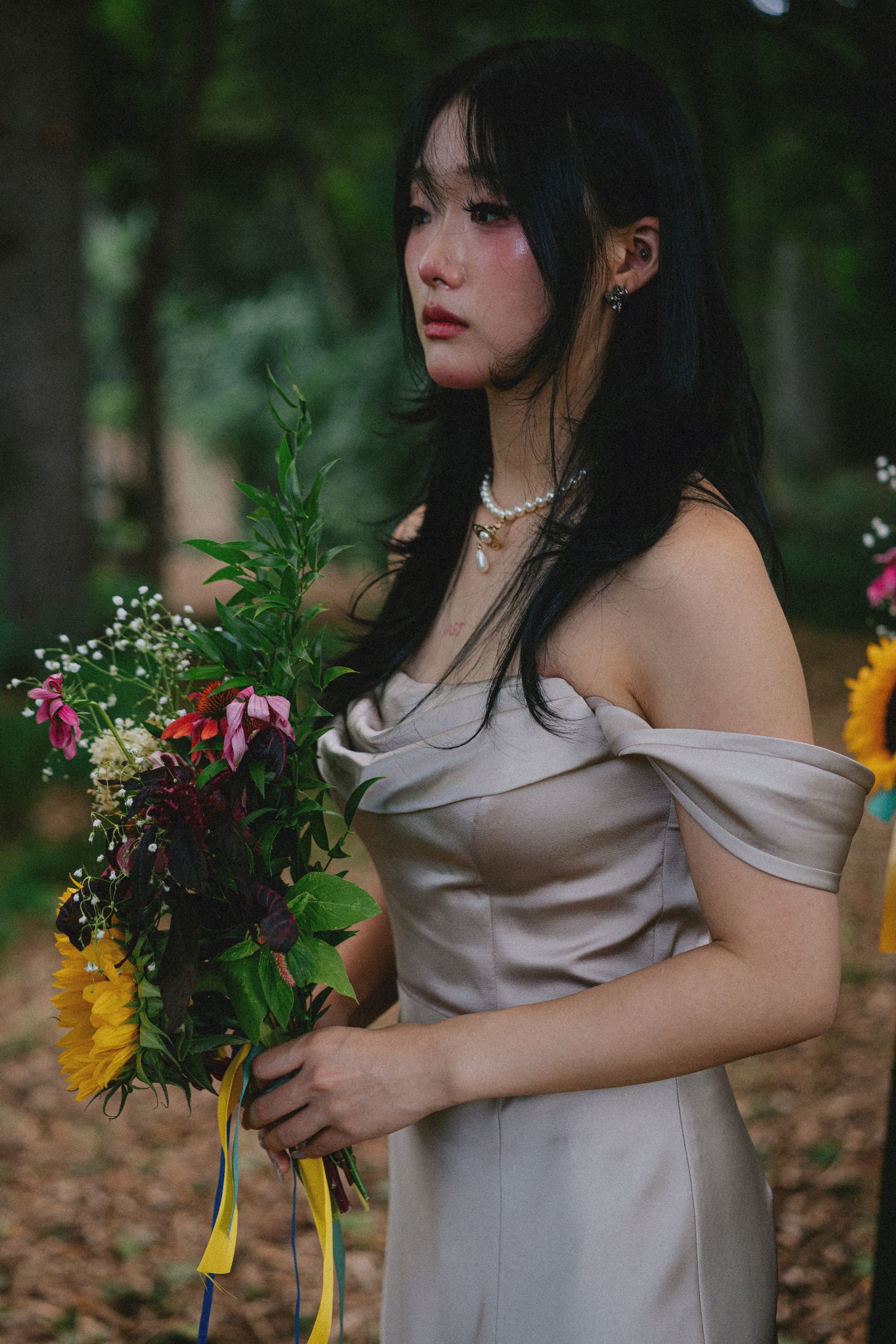 Young Asian woman in off-shoulder gray dress holding colorful flower bouquet outdoors in a lush green forest setting.